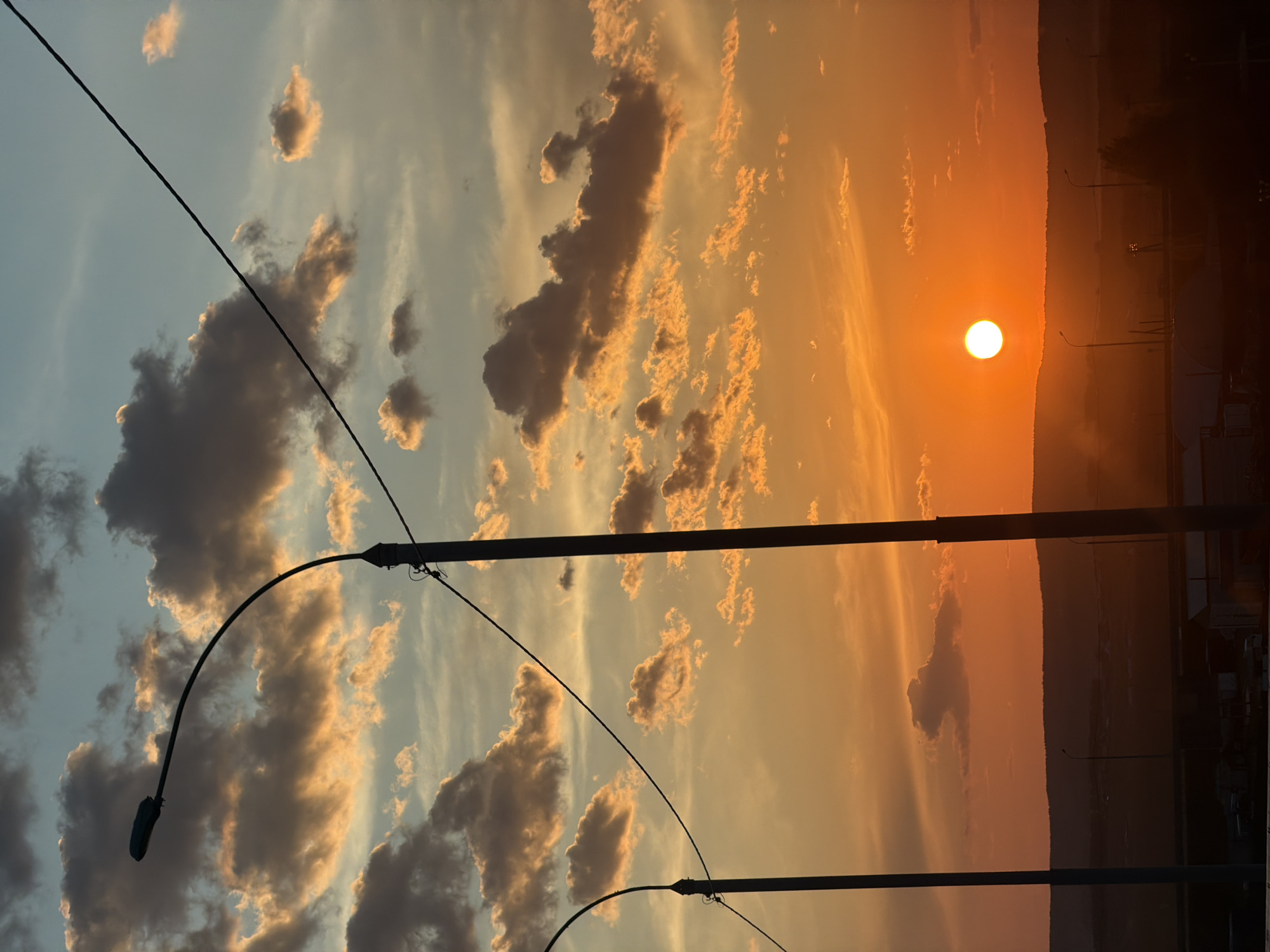 The image captures a sunset scene in what appears to be Krasnoyarsk, Russia, based on visual characteristics. The sky is filled with a mix of colors, ranging from blues and grays in the cloud formations to vibrant oranges and yellows near the horizon where the sun is setting. The sun is a bright, circular orb positioned near a distant treeline or low mountain range.
Several tall light posts with curved arms and attached streetlights are visible in the foreground, silhouetted against the sky. Electrical wires stretch across the scene, intersecting with the light posts.
Below the horizon, there is a darker area, possibly a combination of trees, buildings, and geographical features. The entire scene is oriented in a portrait format, but the image itself is captured horizontally. There are no people visible in the image. The general environment suggests an urban or suburban location with infrastructure.