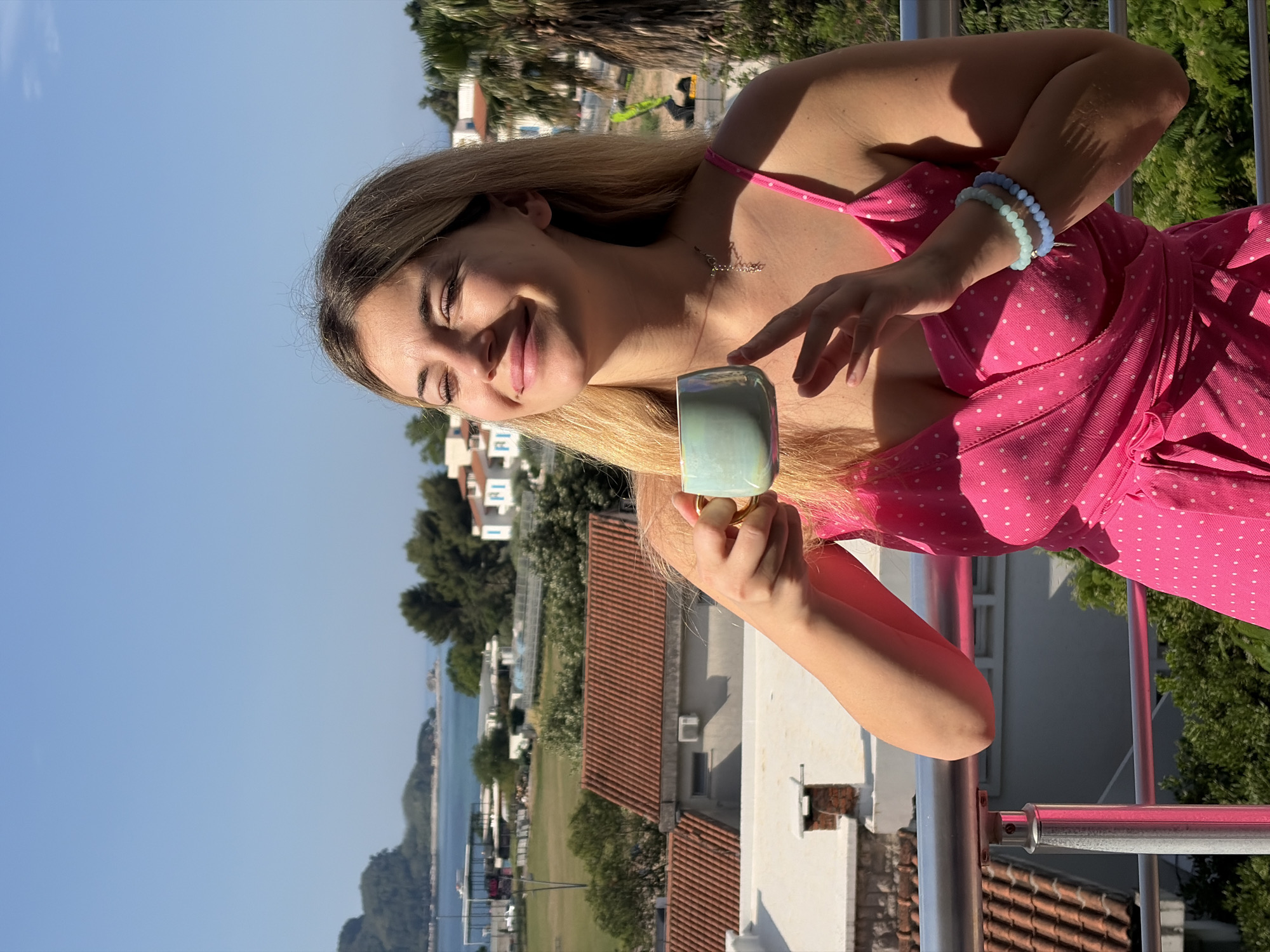 A medium shot depicts a fair-skinned young woman in a pink, polka-dot dress, holding a green mug and looking off-camera with a slight smile. She has blonde hair and wears blue beaded bracelets and a necklace. The image is taken from an elevated position, likely a balcony, overlooking a landscape in Budva, Montenegro. In the background, there's a view of rooftops, trees, and the sea under a clear sky. Some buildings are visible in the distance. The balcony has a metal railing, and the lighting appears to be bright daylight.