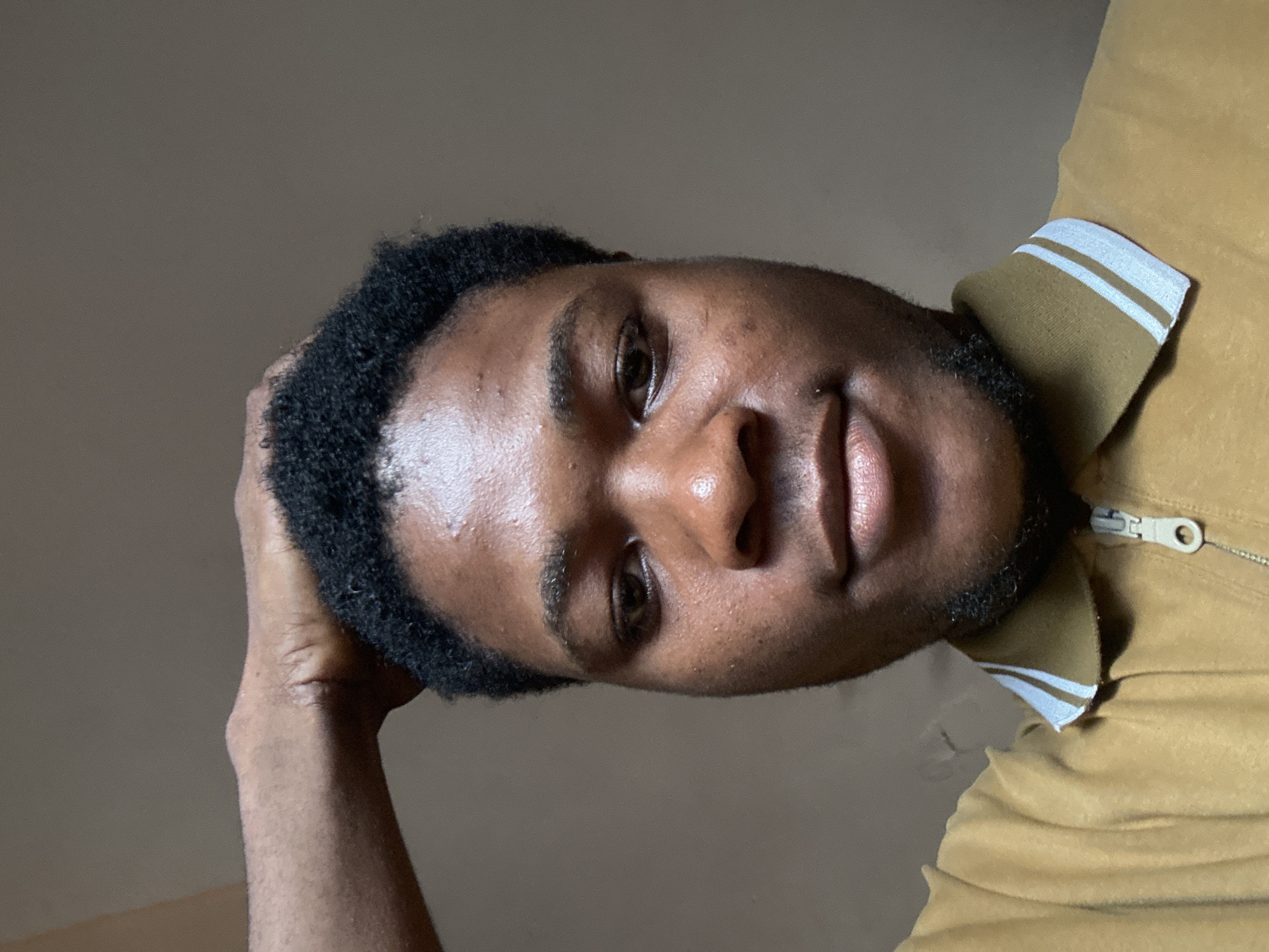 A close-up, slightly angled headshot of a dark-skinned man. He is wearing a gold jacket with white stripes on the collar and a zipper. His left hand is resting on his head. The background is a plain, light brown wall. This image was likely taken indoors. I am unable to determine the location as I have no details in the image to suggest Ihiagwa, Nigeria.