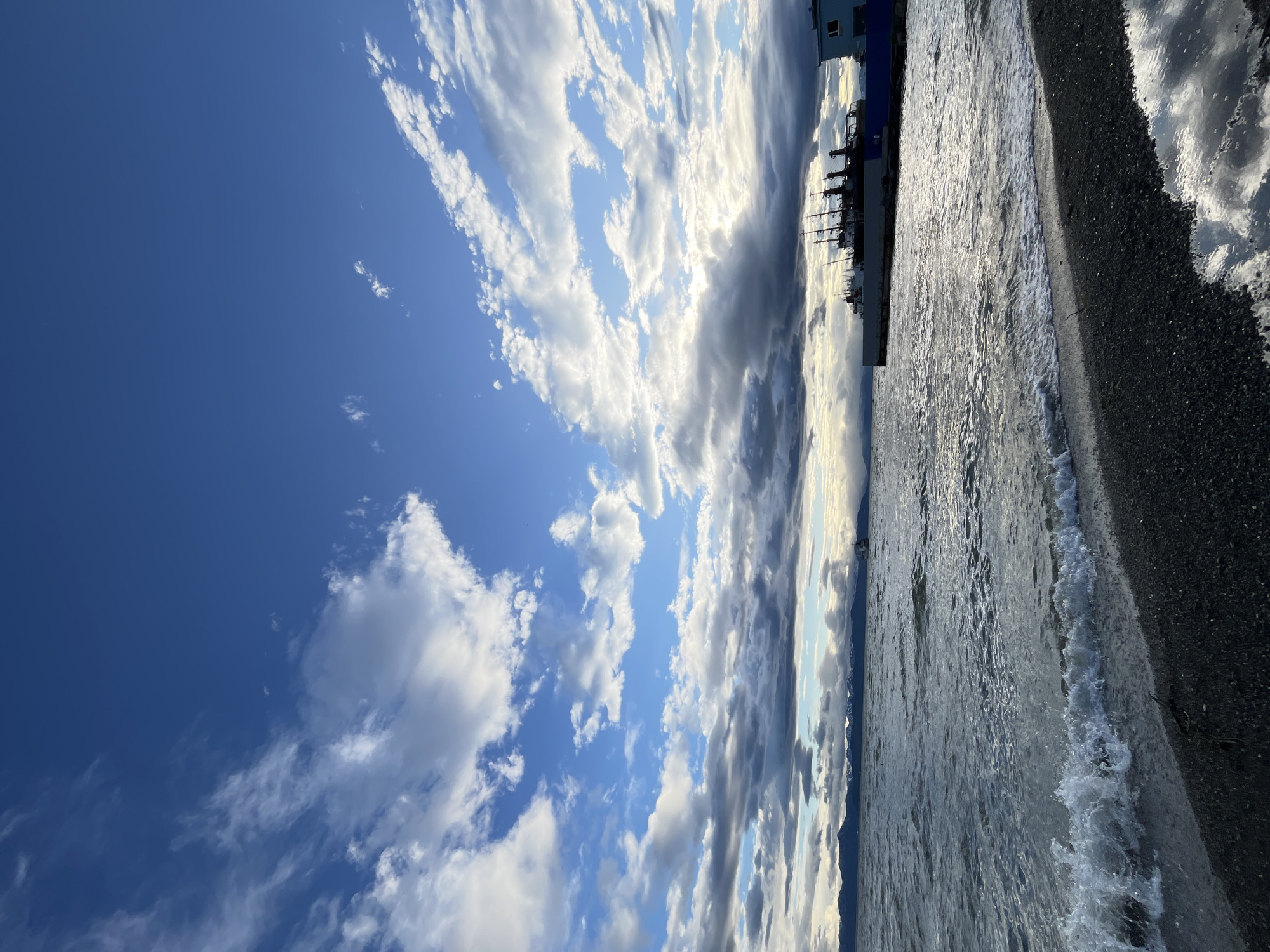 The image depicts a coastal scene, likely within the city of Petropavlovsk-Kamchatsky, Russia, given the characteristic mountainous backdrop and maritime activity. A pebble beach occupies the foreground, with gentle waves lapping the shore. In the midground, the sea stretches towards a distant horizon marked by snow-capped mountains. To the right, a dock or pier is visible, with several docked vessels, including what appears to be a larger ship with multiple masts and a smaller, lighter-colored building or boat structure. The sky above is dynamic, featuring a mix of bright blue patches and dramatic, heavy clouds, with sunlight filtering through in places, creating a sense of approaching or receding weather. No people are discernible in the image.