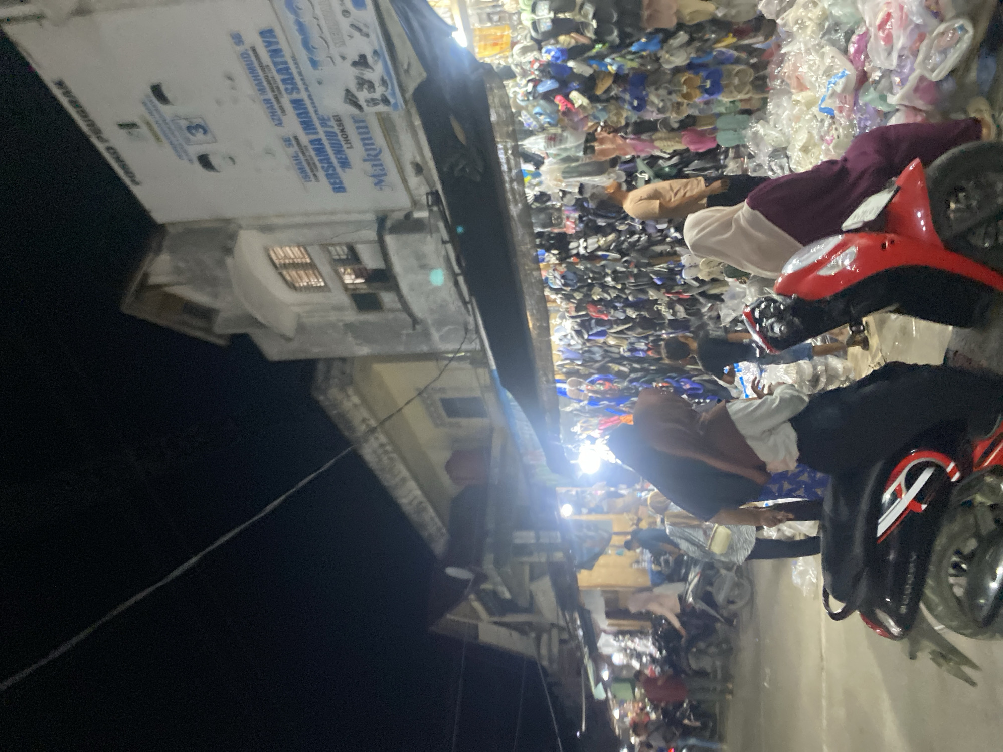The image, taken at night, features a bustling market scene in Lhokseumawe, Indonesia. Piles of goods, including what appears to be footwear and clothing, are spread out for sale. Two individuals are visible near a red motorcycle. Buildings line the street, one of which is labeled with the text "POSKO PEUGRAK" and includes the names "ISMAIL, SE AZHAR MAHMUD, SE" and phrases "BERSAMA IMAM SAATNYA" and "MENUJU PE LHOKSEI Makmur." The environment is lit by streetlights and shop lights. The image is rotated slightly.