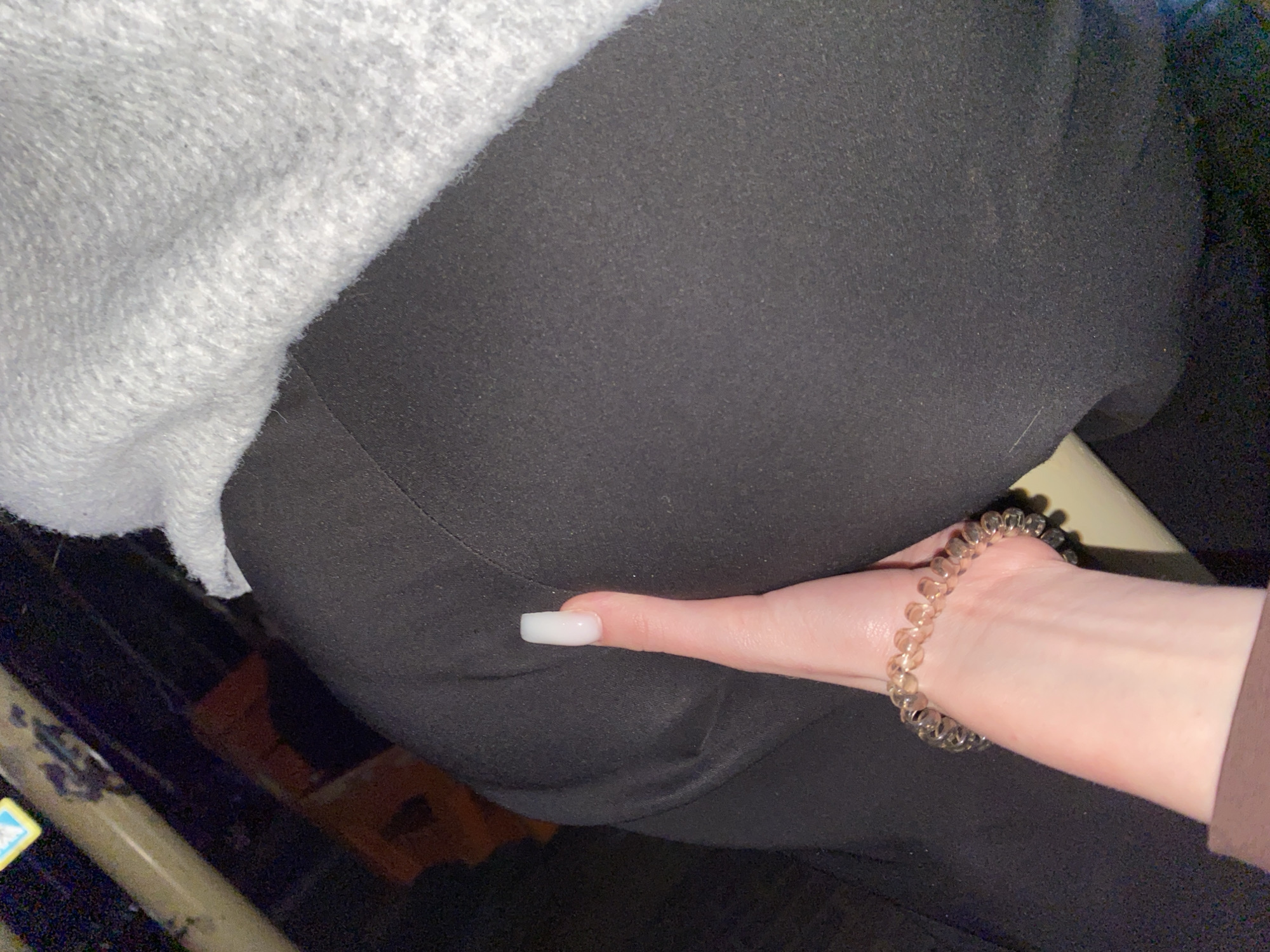 The image captures a close-up view of a person's hand and arm set against a backdrop of clothing. The hand, with light skin, is positioned on a surface of dark gray or black fabric, likely trousers or a skirt. The thumb is extended, featuring a prominent white, square-shaped fingernail. A translucent, coiled bracelet adorns the wrist. In the upper left corner, a section of light gray, textured fabric, possibly a sweater or blanket, is visible. The background is dark and indistinct, with glimpses of dark structures, possibly suggesting an interior setting like a car or public transport. A small, partially visible sticker with yellow and blue elements is present on a dark object in the bottom left corner. The lighting appears dim, contributing to the intimate and somewhat confined feel of the scene. No specific activity is taking place beyond the hand's resting pose.