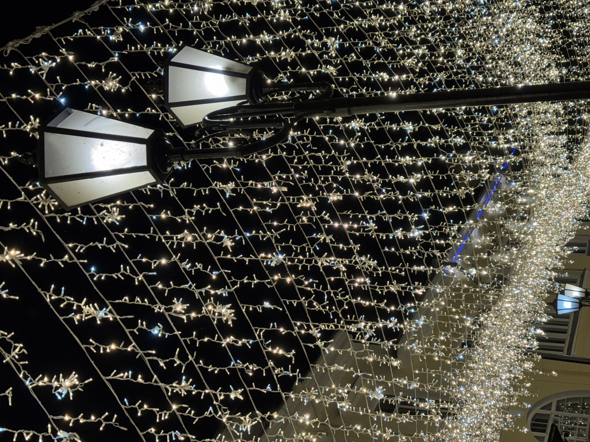 The image depicts a street illuminated by numerous strings of white and blue lights, creating a festive and wintry atmosphere. Two large, hexagonal street lamps with frosted glass panels are prominently featured, emitting a bright, warm light. The lamps are mounted on a black, ornate pole. The strings of lights hang in dense curtains across the width of the street, with some lights appearing as small, star-shaped clusters. The background is dark, suggesting nighttime. In the lower left portion of the image, parts of a yellow building with arched windows are visible, hinting at an urban setting. The scene is likely a street in Vitsebsk, Belarus, decorated for a holiday or special event. There are no people or animals visible in the image.