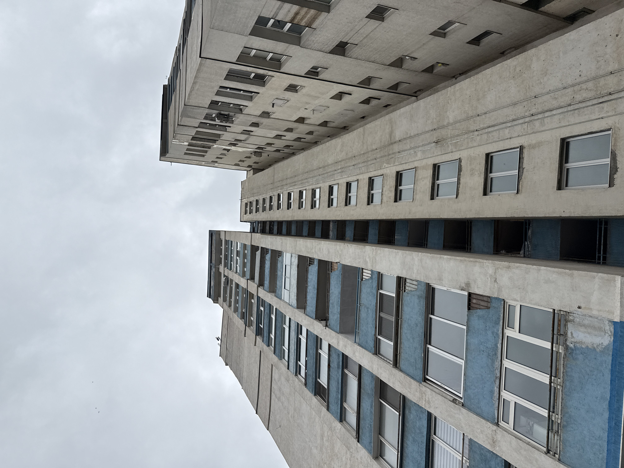 An upward-angled shot captures two adjacent concrete buildings against a cloudy sky. The building on the right extends vertically and has a repetitive pattern of windows, some of which have blue painted sections between the concrete frames. The building on the left is constructed from a similar concrete material but has smaller, square windows. The architectural style appears to be utilitarian, with a minimalist design. The location is possibly in the city of Sofia, Bulgaria.