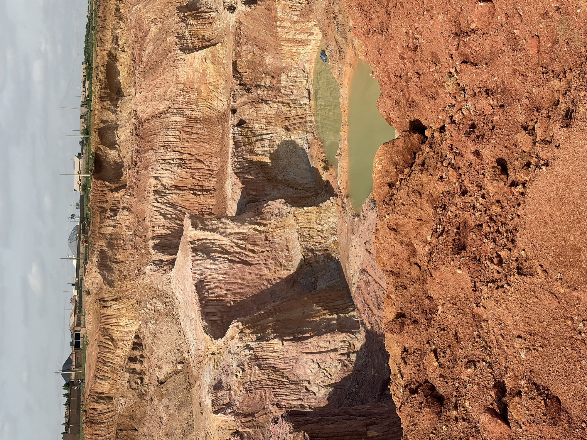 This image captures an active quarry site, likely for clay extraction, in Kwang, Nigeria. The foreground is dominated by a heap of reddish-brown soil and rocks, indicative of excavated material. The midground features a deep excavation with sheer, layered walls of soil in shades of red, orange, and yellow, revealing distinct geological strata. Two pools of murky, light green water have formed at the bottom of the quarry. A dark-colored vehicle, possibly a pickup truck or utility vehicle, is parked near the edge of the larger pool. In the background, the quarry edge meets the landscape, where several residential buildings and power lines are visible. A few individuals are present on the upper edge of the quarry, appearing to observe the activity or work in the area. The sky is overcast with scattered clouds. The overall scene suggests ongoing industrial activity within a developed or developing urban periphery.