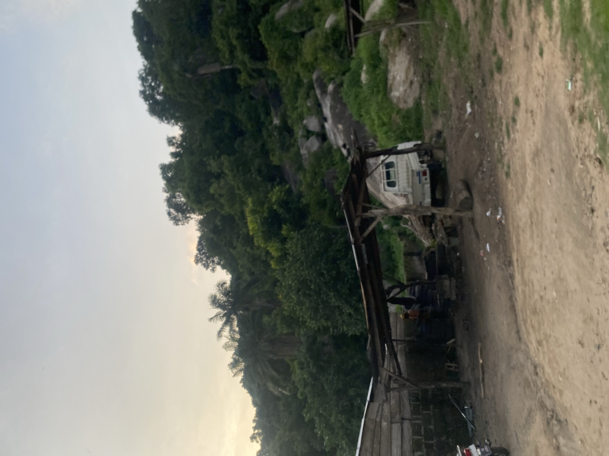This image captures a semi-rural landscape in Enwan, Nigeria, under a soft, overcast sky. The background is dominated by a densely vegetated hill covered with various green trees and shrubs, including noticeable palm trees. Large grey boulders are interspersed throughout the hillside, protruding from the foliage.
In the midground, a collection of rudimentary structures is visible on uneven dirt ground. To the left, a building constructed from wooden planks and possibly concrete blocks features a light-colored roof. Central to the scene is an open-sided shelter with a flat, dark roof supported by wooden poles and a rough-hewn wooden framework. Beneath this central shelter, one individual wearing an orange/yellow top is partially visible, appearing to stand near stacked dark objects, likely tires. A small, white utility truck, possibly a mini-pickup, is parked beneath this same shelter, facing away from the viewer. To the far right, a smaller, similar open-sided shelter with a dark roof is partially visible.
On the far left, a portion of a white and blue motorcycle is parked next to the wooden plank building. The foreground consists of light brown, unpaved dirt with scattered patches of green grass, particularly to the right. Various pieces of light-colored litter and debris are dispersed across the ground. A segment of a tree log lies on the dirt near the white utility truck. The overall lighting is soft and diffused, with the sky transitioning from pale blue overhead to a faint yellowish hue near the horizon on the left.