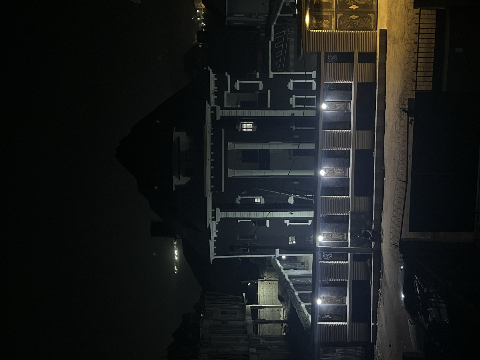 The image captures a tall, multi-story building at night in Abuja, Nigeria. The main structure, oriented vertically, dominates the left side of the frame, showcasing a dark facade contrasted with prominent white architectural details such as columns, window frames, and cornices across its numerous levels. Several windows reveal interior lights, while numerous exterior lights illuminate the building's facade and balconies, casting bright glows and creating deep shadows.
To the right of the main building, a boundary wall or fence is visible, sections of which are topped with barbed wire, suggesting security measures. Further right, an ornate metal gate with intricate decorative patterns stands, likely marking an entrance to the property. A dark-colored vehicle is partially visible parked near this gate on what appears to be a street or driveway.
Towards the bottom left, at the base of the main building, less finished structures are evident, possibly indicating ongoing construction or ancillary buildings. These sections show exposed concrete blocks and scaffolding-like elements, also illuminated by artificial lights. The sky is entirely dark, confirming the late-night setting. No people or specific activities are discernible in this static scene. The bright lighting suggests occupancy and a focus on security within an urban environment. No visible text is present.