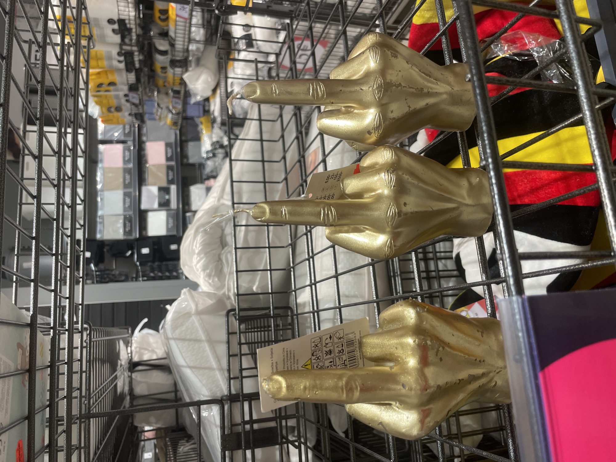 Three gold-colored candles shaped like middle fingers are displayed on a metal shelf in a retail store. The candles are positioned in a row from left to right, with the leftmost candle partially obscured by a product tag. The middle and rightmost candles are fully visible. Behind the candles, various products are stacked on shelves, including what appear to be bedding or fabric items wrapped in plastic. The background shows more shelves stocked with packaged goods, some in yellow and white packaging, and others in muted tones. The overall environment is a retail display, likely in a department store or home goods store. The provided hints suggest the location is Iserlohn, Germany, though this cannot be visually confirmed from the image alone.