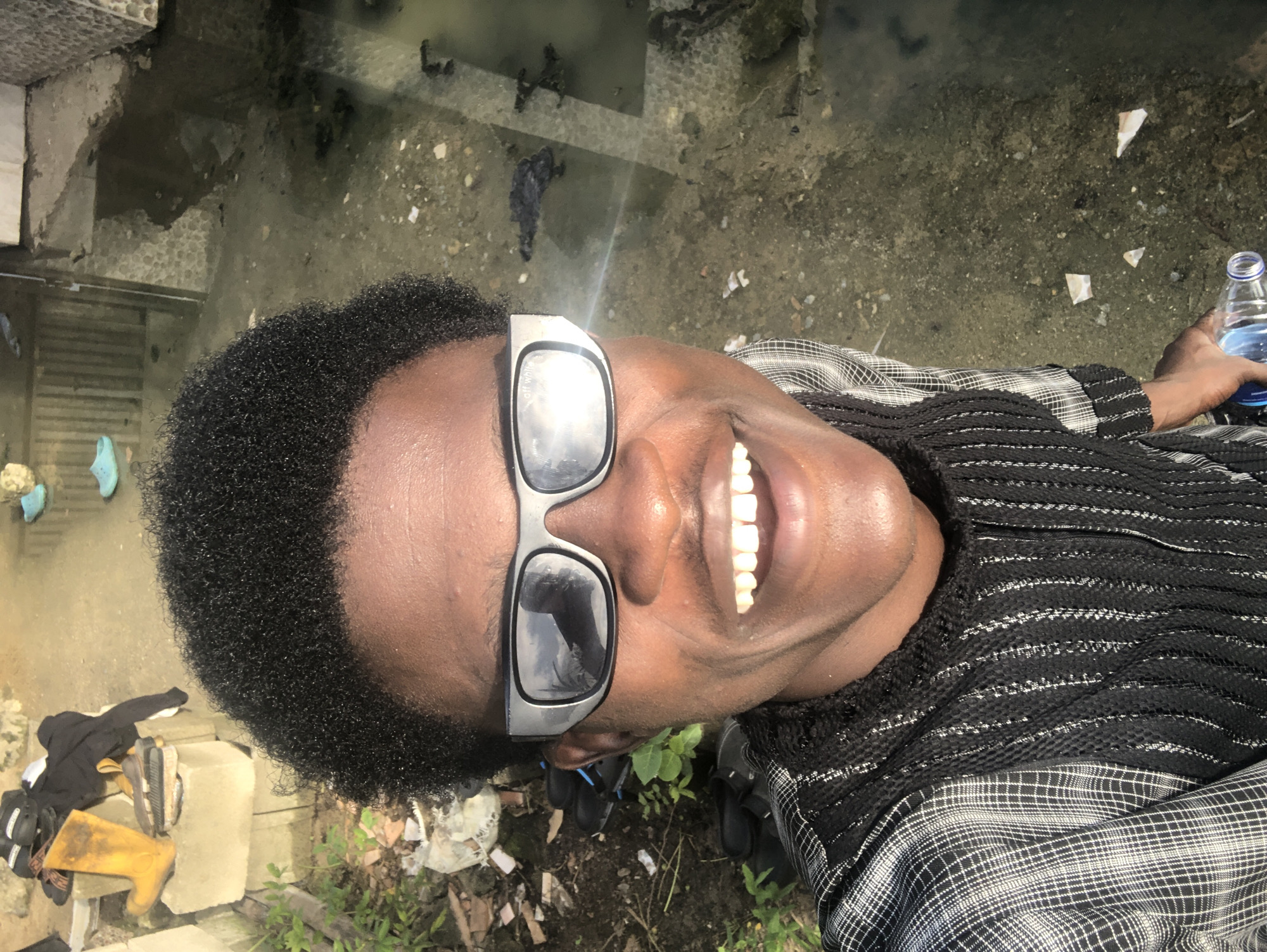 A close-up, high-angle selfie captures a young Black individual from the chest up, facing the camera with a wide smile. They are wearing futuristic-style silver-rimmed sunglasses and a patterned shirt with black and white checks on the sleeves and a textured black knit fabric on the chest. The background appears to be an outdoor area in Ado, Nigeria, with a muddy, water-filled ground that reflects the environment. To the left, there are piles of construction materials, including cement blocks, and various discarded items such as yellow boots, sandals, and black clothing. Green foliage sprouts from the ground on the left side. The overall impression is one of an urban or semi-urban environment that is somewhat unkempt.