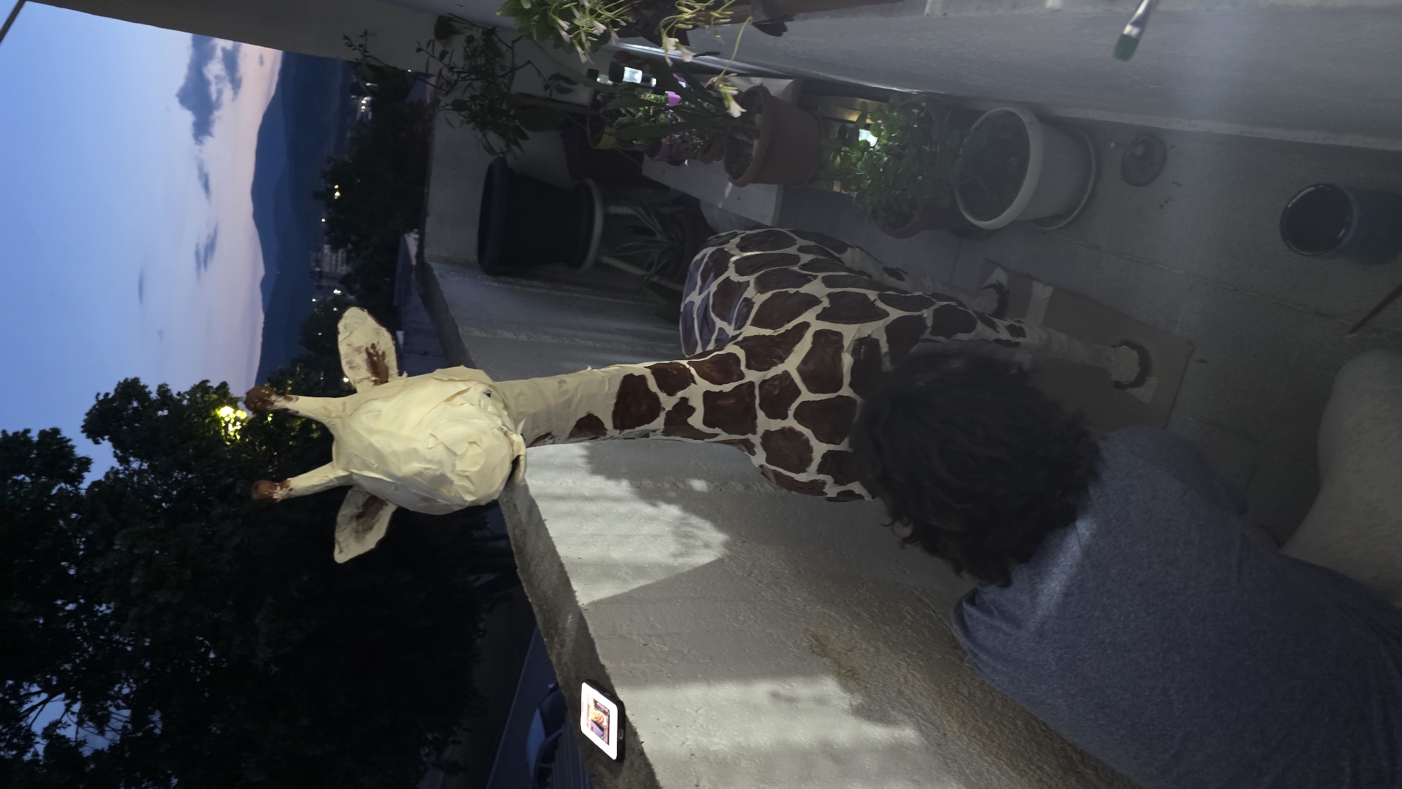 The image shows a person in a blue shirt working on a large, partly finished giraffe sculpture on an outdoor balcony. The giraffe's head is covered in a white material, likely paper mache, and its body is painted with brown spots. Several potted plants line the balcony. In the background, there is a view of a city with mountains in the distance under a dusky sky. The scene is likely in Blagoevgrad, Bulgaria, given the urban setting and the presence of mountains similar to those found in the region. There is a picture frame on the balcony wall, and painting supplies are scattered around.