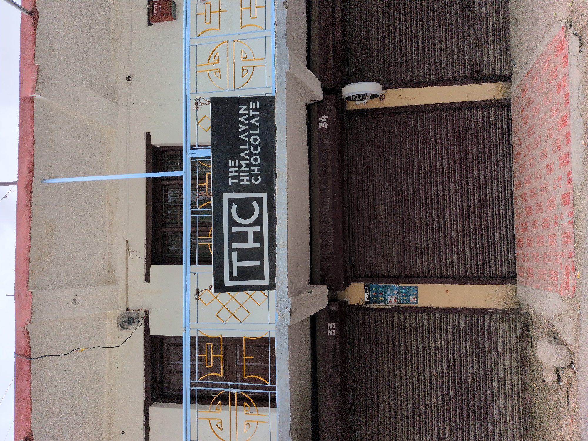 The image captures the facade of a two-story building in Leh, India, with a focus on the entrance to "THE HIMALAYAN CHOCOLATE" shop. The shop is identified by a black sign with white lettering that reads "THC THE HIMALAYAN CHOCOLATE". Above the shop, a blue railing with yellow decorative elements adorns a balcony. Two wooden doors are visible on the upper level, each with a grid-like pattern. A utility box is mounted on the building's exterior on the left, and a red "LETTER BOX" is visible on the right.
The ground level features two large, dark brown metal roll-up shutters, suggesting commercial establishments. The shutter on the left is marked with the number "33", and the shutter on the right is marked with "34". A vertical beige pillar separates these two shutters. A poster, possibly an advertisement or information leaflet, is affixed to the pillar between the shutters. A white circular object, possibly a security camera or a light fixture, is attached to the pillar above the poster. The ground in front of the shutters is a mix of dirt, rocks, and red patterned paving stones. The overall scene is one of a commercial street in Leh, India, under an overcast sky.