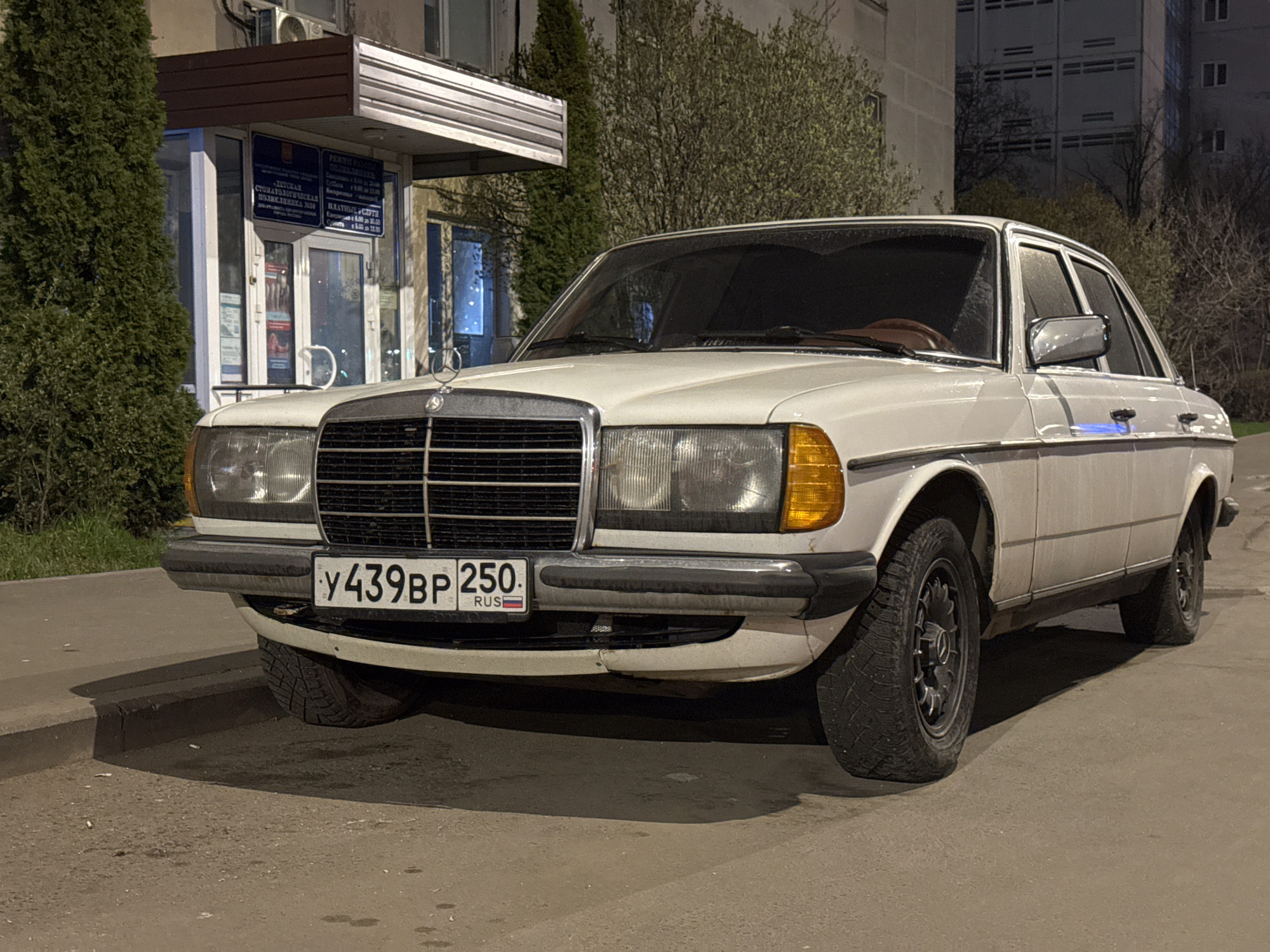 A white Mercedes-Benz W123 sedan with the Russian license plate "У439ВР 250 RUS" is parked in what appears to be an urban setting, possibly in Moscow, Russia. The car is positioned in front of a low building which looks like a dental clinic with signs in Russian. The lighting suggests it is either dusk or night. The car appears to be somewhat older, with visible dirt or wear on the body. The surrounding environment includes trees and multi-story buildings, characteristic of a city. The curb is visible along the edge of the road. No people are clearly visible in the frame.