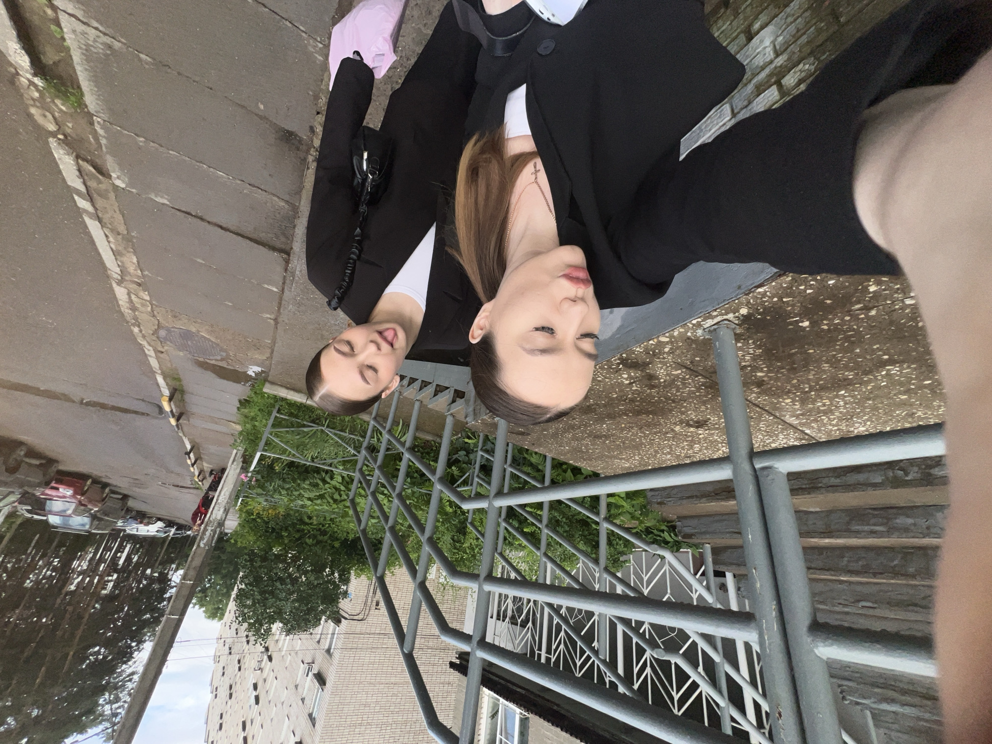 Two young women are taking a selfie outdoors in the city of Chaikovsky, Russia. They are standing on a paved walkway next to a set of concrete stairs with metal railings. The woman in the foreground is wearing a black blazer over a white top and a cross necklace. She is puckering her lips for the camera. The woman behind her is also wearing a black blazer over a white top and is sticking her tongue out. She has a small black purse on her shoulder and is holding a pink plastic bag. In the background, there is a brick apartment building, lush green foliage, and a street with parked cars and a telephone pole. The ground appears wet, suggesting it might have rained recently.