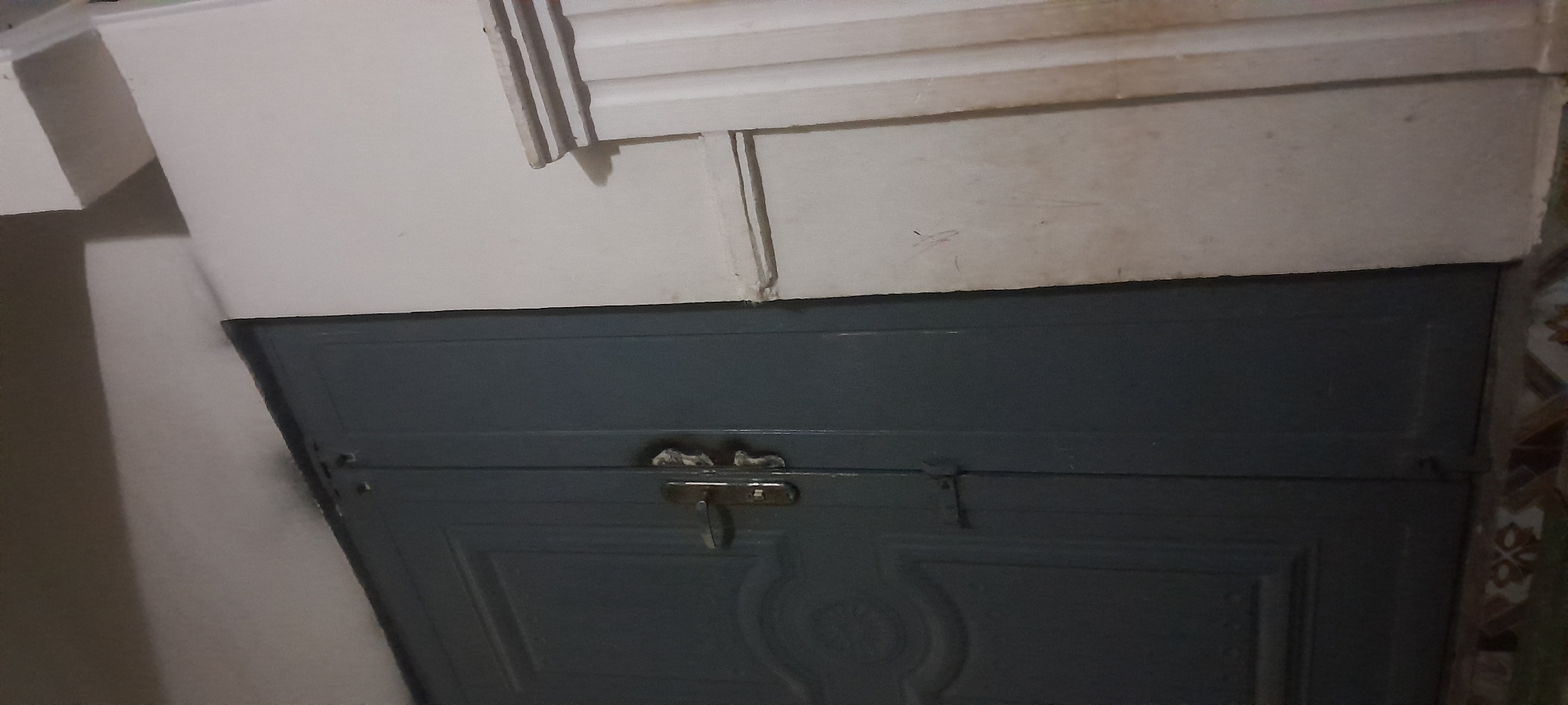 A medium shot, taken from a low angle, shows the exterior of a dark gray double door with ornate paneling on the left door. The doors are closed. A silver-toned door handle with a keyhole is mounted vertically on the left door, and a small, dark metal latch is visible near the center seam of the doors. To the right of the door is a section of a white wall, which has a decorative, fluted column or trim piece running vertically. The wall has some scuff marks and faint lines. The ceiling above the door appears to be a light color and is casting a shadow onto the top of the door frame and the wall. The bottom of the image shows a decorative tile border on the floor. The lighting is dim, creating shadows.