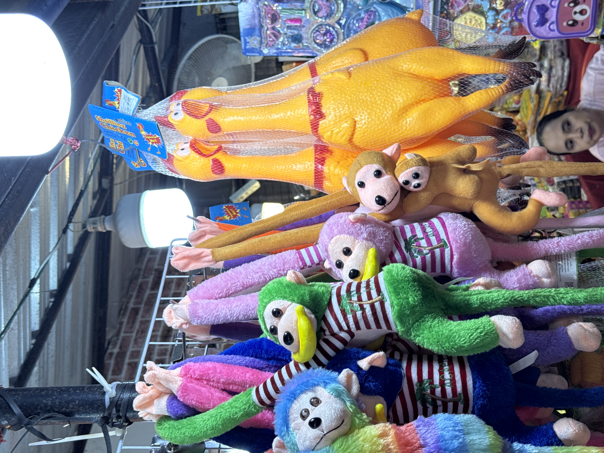 In Karon, Thailand, a stall displays a variety of colorful novelty toys. In the foreground, several stuffed monkeys with large yellow beaks and striped shirts hang from a rack. A blue-headed monkey with rainbow-striped arms is on the left. To its right, a green-headed monkey wears a red and white striped shirt with a palm tree graphic and the word "ACAPULCO" stitched below. A purple-headed monkey with a similar shirt and graphic is also visible. Behind the monkeys, a net bag contains several large, bright yellow rubber chickens with red accents around their necks. A woman is partially visible in the lower right corner, looking towards the left. In the background, a white oscillating fan is mounted on a wall, and various other items are displayed on shelves. The stall appears to be outdoors, under a corrugated metal roof supported by metal beams.