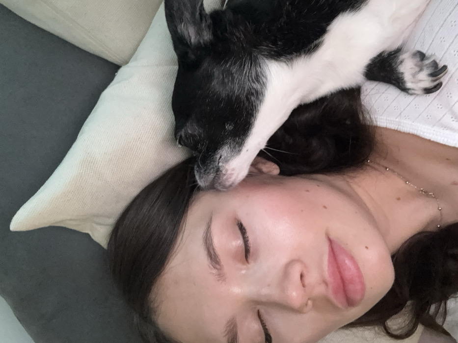 A woman lies on a gray sofa with a white pillow under her head. She has dark hair and her eyes are closed. A black and white dog is lying on her head, its face nuzzled against her forehead. The woman is wearing a white top and a delicate necklace. The scene is intimate and peaceful, capturing a moment of affection between the woman and her pet.