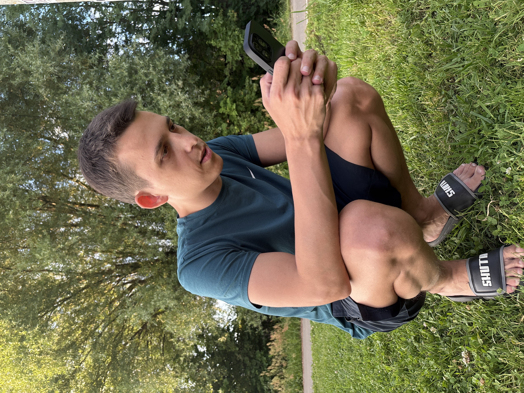 A young man is sitting on the grass, holding a smartphone and looking at it. He is wearing a dark teal t-shirt and dark shorts. The scene is outdoors, with green grass and trees in the background. The lighting suggests it is daytime, likely late afternoon given the angle of the sun casting shadows. The man appears relaxed, engrossed in his phone. The setting seems to be a park or a grassy area in a residential neighborhood. The text "SHUT!" is visible on his sandals.
