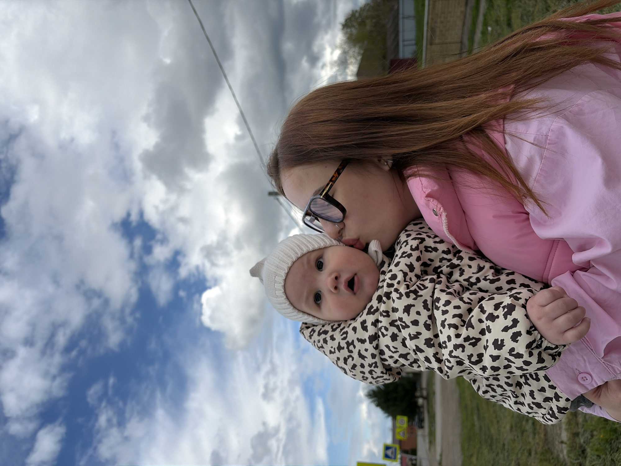 A woman is kissing a baby in Novopokrovskaya, Russia. The woman has long brown hair and is wearing glasses and a pink jacket. The baby is wearing a white hat and a beige leopard-print jacket. They are outside, and the sky is cloudy. Traffic signs are visible in the background.