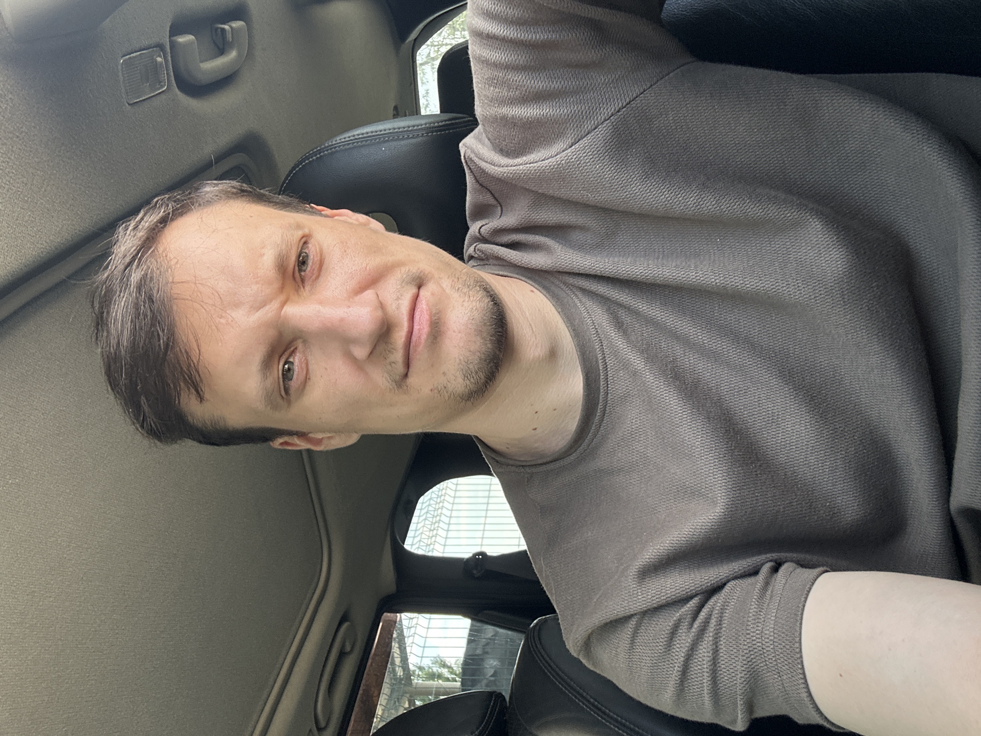 A selfie of a man taken inside a car. He is in the driver's seat, and the camera is positioned slightly above eye level, looking down at him. The man has short, dark hair and is wearing a brown t-shirt. He has a faint stubble on his chin and his mouth is slightly open. The car's interior is visible, including the ceiling, a sunroof, and the back of the driver's seat. The windows show an outdoor scene with a fence and some greenery. The image was taken in Shatava, Ukraine.