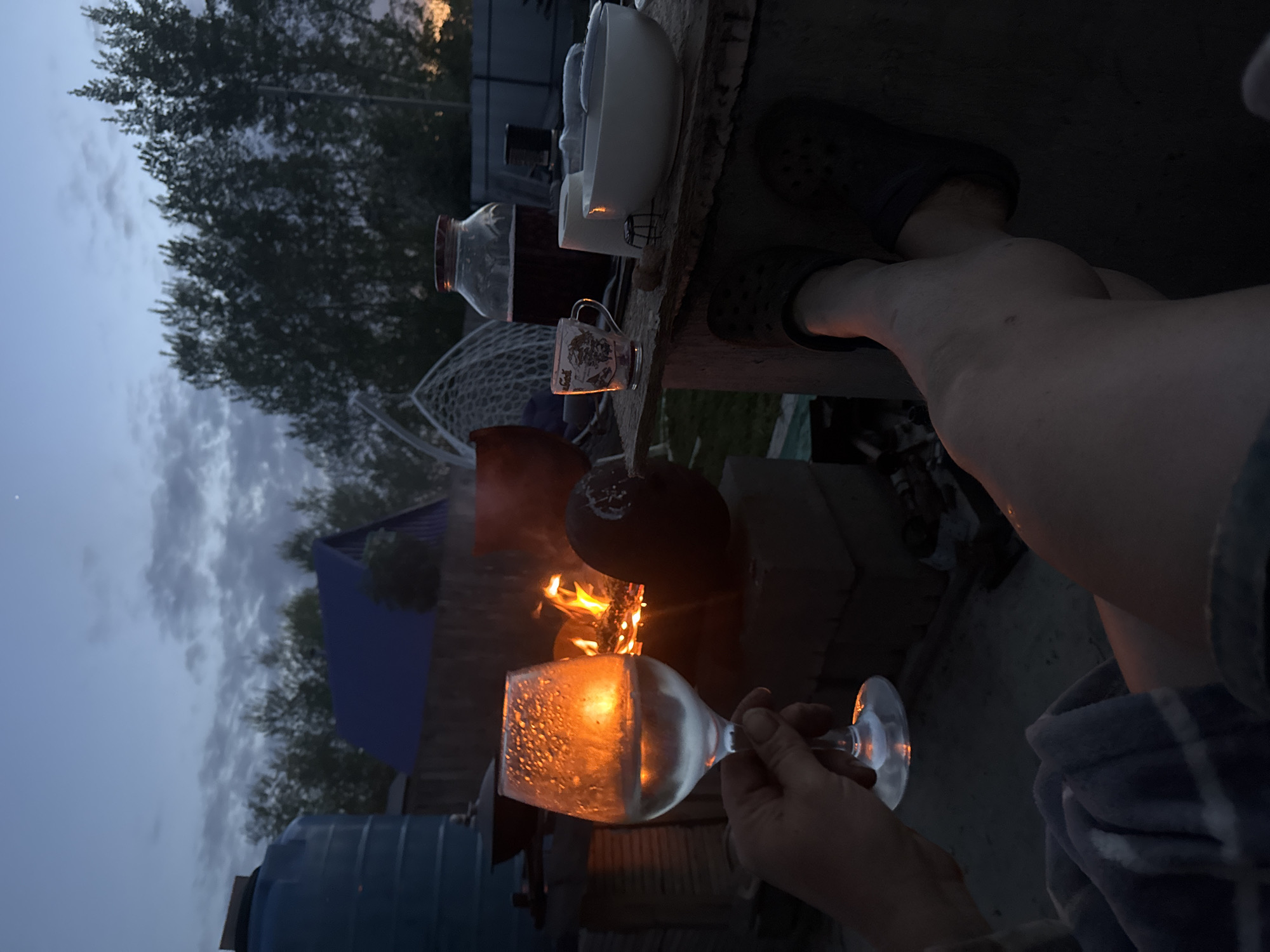 The image depicts a person relaxing near a fire outdoors, possibly in a backyard setting. The location appears to be near Chaikovsky, Russia, based on metadata. The person is wearing slides and is seated, holding a wine glass that is reflecting the light from the fire. In the background, there is a fire burning in what appears to be a makeshift fire pit made of bricks. Beside the fire pit is a blue water tank. There is a table with various items on it, including a jar, cups, and bowls. The sky is overcast and it appears to be dusk or evening. In the background are trees and the roof of a house.