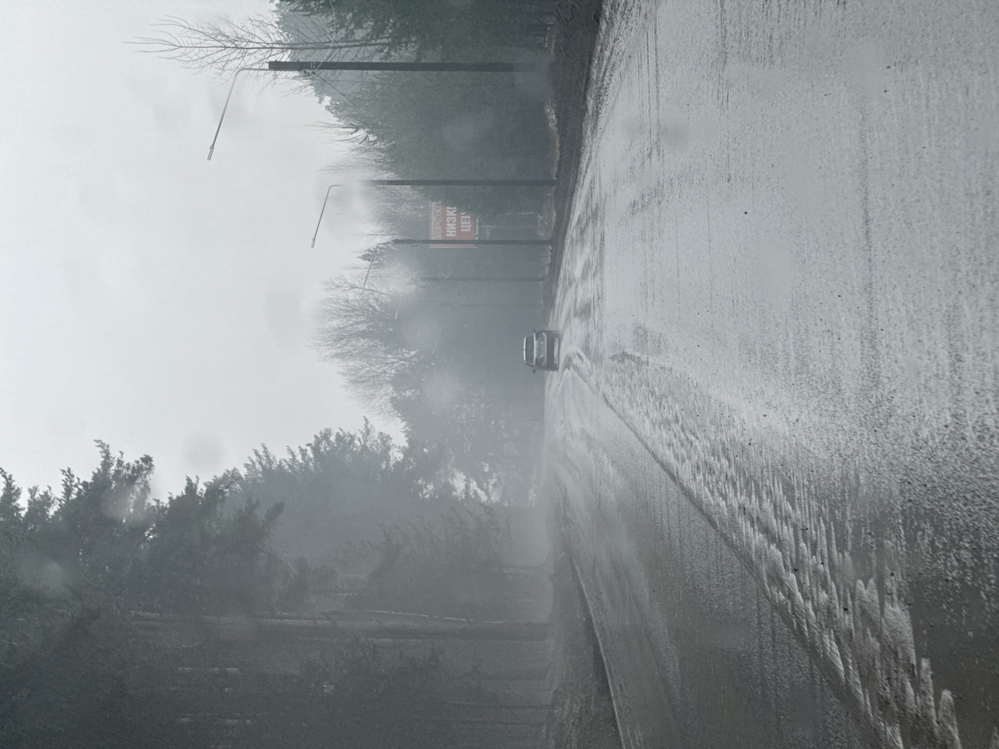 The image depicts a wet road, likely asphalt, running through what appears to be a wooded area in heavy fog or rain. The road surface shows signs of water accumulation. Trees and some utility poles, including one with a street lamp, are visible along the road's edge. Further down the road, a sign is barely visible. A car is driving on the road. This photo was taken in the city of Chaikovsky, Russia.