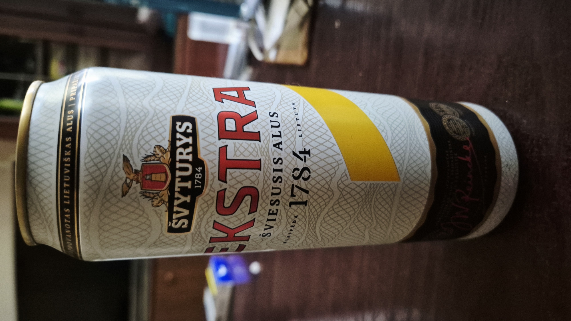 A close-up, eye-level shot shows a tall, slim can of Švyturys Extra beer, predominantly white with a gold rim. The can features intricate, light grey cross-hatching patterns. Prominently displayed is the Švyturys logo, a shield with a lighthouse and eagle emblem, above the text "ŠVYTURYS 1784". Below this, the word "EKSTRA" is written in large, red, sans-serif capital letters. Further down, smaller text reads "ŠVIESUSIS ALUS", followed by "KLAIPĖDA 1784 LIETUVA" and a yellow diagonal band. The bottom of the can has a dark label with ornate gold detailing and what appears to be a handwritten signature in pink. The can rests on a dark, polished wooden surface, likely a table. The background is blurred, showing hints of furniture and a dark television screen on the left. The overall lighting is diffused. No people are visible in the image. The scene does not provide any specific clues about its location within the city of Varena, Lithuania.