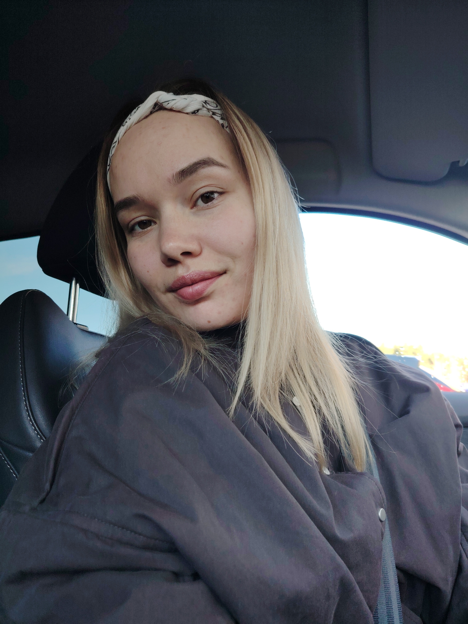 A fair-skinned young woman with blonde hair and a patterned headband is seated in what appears to be a car. She is wearing a dark-colored jacket or coat with visible buttons and a seatbelt. The background visible through the car window suggests an outdoor environment. The image provides insufficient data to confirm the location as Markovskii, Russia.
