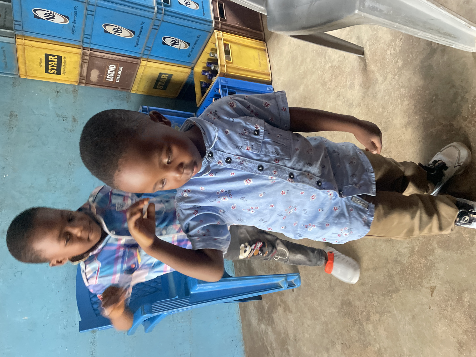 The image depicts two young boys in what appears to be a storage area. The boy in the foreground, wearing a light blue patterned short-sleeved shirt and tan pants, is looking down. He has short dark hair and is standing on a concrete floor. Behind him, another boy, wearing a blue and pink plaid shirt, is seated on a blue plastic chair and looking down and to his left, with his right arm raised.
The environment shows stacked crates, primarily blue and yellow, with some bearing labels such as "STAR" and "LEGEND EXTRA STOUT". Blue plastic crates containing bottles are visible, as is a grey plastic chair in the foreground. The wall behind the boys is painted light blue. The lighting suggests an indoor setting, possibly a shop or a warehouse. The presence of "Nigerian Breweries Plc" branding on some crates indicates the location is likely Nigeria, and given the prompt's context, this scene could be in the city of Jos, Nigeria.