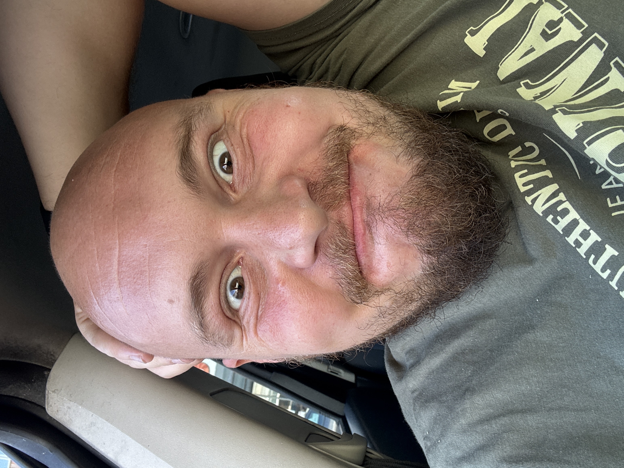 A selfie taken from inside a vehicle shows a bald man with a full beard looking at the camera. He is wearing a green t-shirt. His left hand is touching the top of his head. The background is blurred but suggests the interior of a car, with a black seat visible on the left. The image is taken in daylight. There is no information in the image to suggest a location in the city of Cork, Ireland.