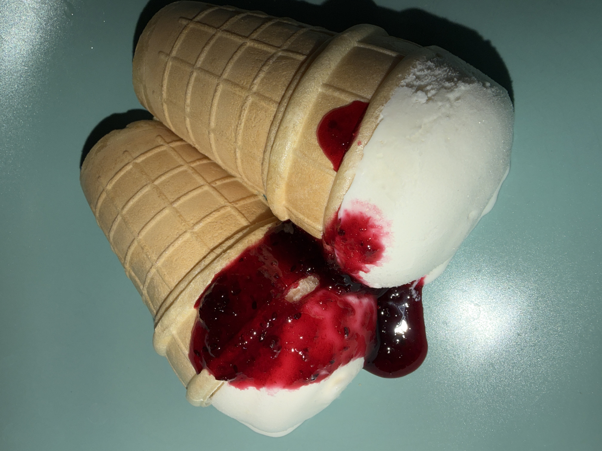 The photograph displays two waffle ice cream cones resting on a textured, light blue-green surface. Both cones contain white ice cream, which is visibly melting. A rich, dark red berry jam or sauce, featuring small fruit pieces or seeds, oozes from the cones and pools on the surface beneath them. The waffle cones exhibit a characteristic grid pattern, and the white ice cream shows some frosty crystallization on its surface.
Artificial lighting illuminates the scene from the upper right, casting defined dark shadows behind the cones and creating bright specular highlights on the glossy berry sauce and the subtly reflective, speckled surface. No people are visible, nor is any text present in the image. The melting state of the ice cream suggests it has been left out for some time or has fallen, creating a somewhat messy yet appealing visual. This scene was captured in Chaikovsky, Russia.