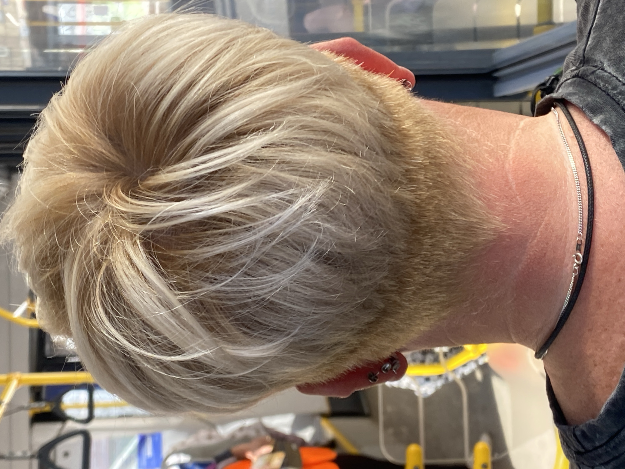 This image shows the back of a person's head with short, blonde hair. The hair is styled in layers, with a textured cut at the nape of the neck. The person is wearing earrings in both ears, and a black cord necklace with a silver chain. The background appears to be the interior of a public transport vehicle, possibly a bus or train, with yellow poles and handles visible. The lighting suggests it is daytime. The image does not provide any information that would allow for identification of the location as Novokuznetsk, Russia.