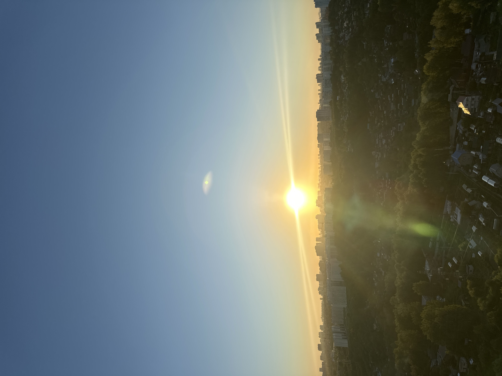 The image depicts a sunrise over a city skyline, likely Novosibirsk, Russia, given the architectural style and vegetation. The sun, a bright orb, is positioned near the horizon, casting a warm, golden glow across the sky and illuminating the city in the distance. The sky transitions from a deep blue at the top to a lighter hue near the sun, with some atmospheric haze present.
In the foreground, a densely populated area with numerous small houses, some with greenhouses and gardens, is visible. This suggests a suburban or dacha settlement. Trees and vegetation are abundant, indicating a green and possibly rural or semi-rural environment bordering the city. Power lines and some infrastructure are also discernible within this area.
Further in the background, a more modern cityscape unfolds, characterized by numerous apartment buildings and taller structures. These buildings appear to be of Soviet-era or contemporary Russian design, consistent with urban development in cities like Novosibirsk. The sunlight creates lens flares and lens dust artifacts, with a prominent green flare extending downwards from the sun. No people or animals are visible in the frame. The overall scene captures a tranquil morning atmosphere as the city awakens.