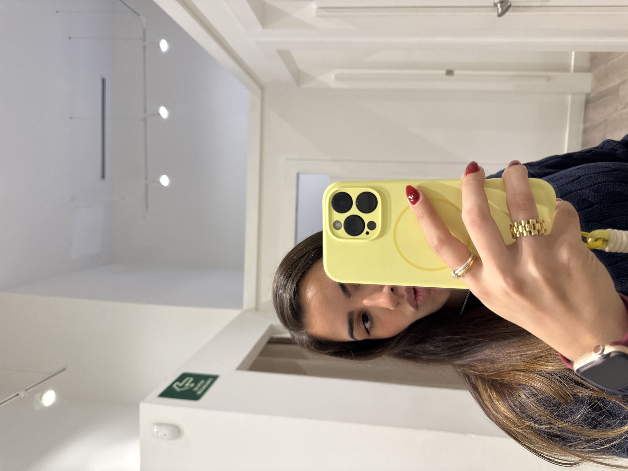 A woman is holding a yellow cell phone up to take a selfie. She is indoors in a room with white walls, a white door, and white trim. There are lights on the ceiling. The woman is wearing rings on her fingers and a watch on her wrist. It is not possible to discern from this image whether the location is San Francisco Ocotlán (Ocotlán), Mexico.