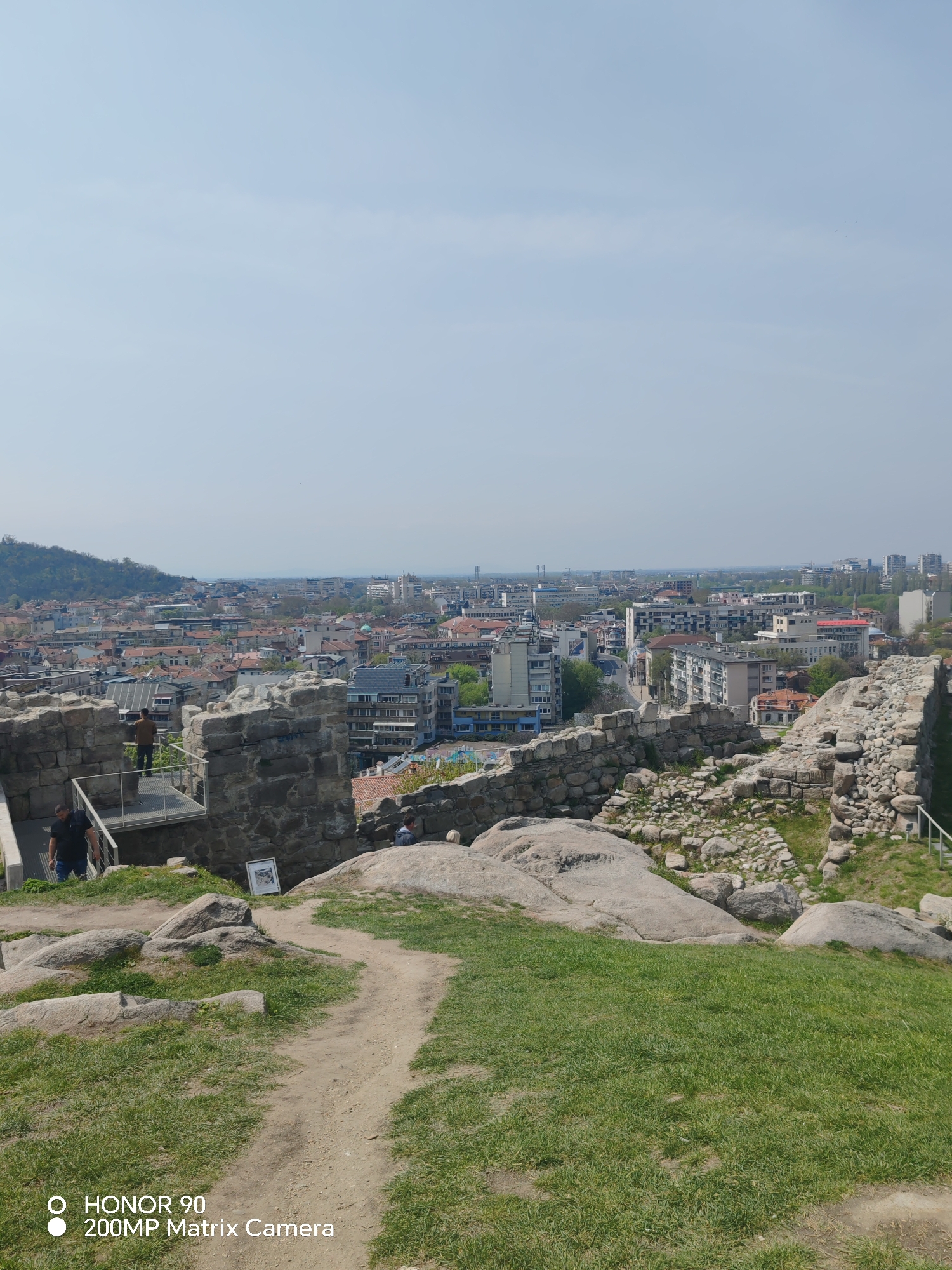 The image depicts a view from Nebet Tepe in Plovdiv, Bulgaria, overlooking the city. In the foreground is a grassy area with a dirt path leading toward stone ruins. The ruins consist of fragmented stone walls and rocky outcrops. Two people are visible near the ruins: one is walking down a set of stairs with a metal railing, and another is standing further back on a metal viewing platform. A third person is seen sitting on some rocks. The middle ground features a cityscape with buildings of various sizes and colors, predominantly with red-tiled roofs. The background shows a pale blue sky.