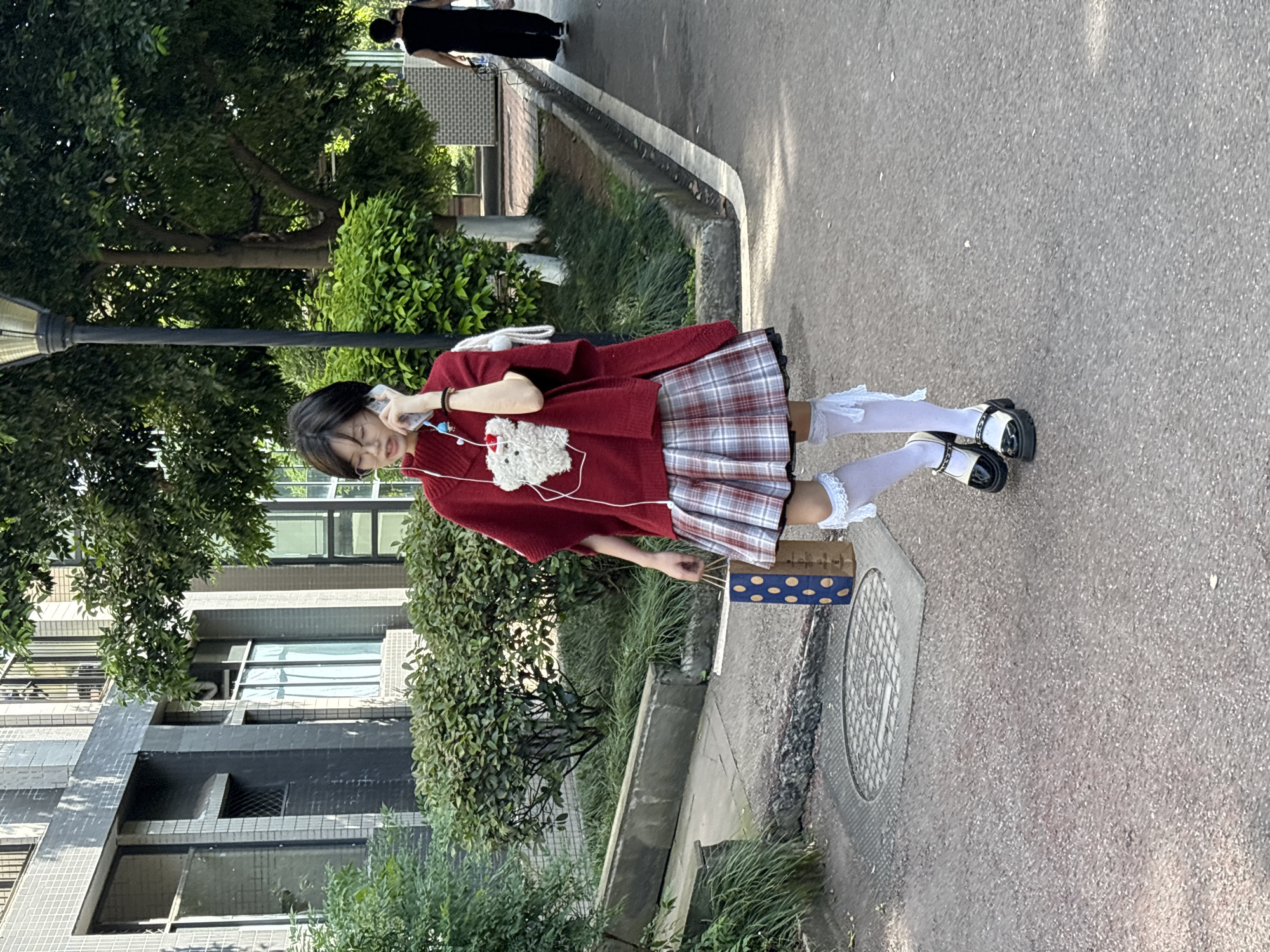 A young female is captured walking on a paved path outdoors in Jiangjin, China, during bright daylight. She is the primary subject, with short dark hair and eyeglasses, holding a smartphone to her left ear with white wired earbuds. She wears a loose-fitting red knitted sweater, which has a fluffy white bear-like patch on the front, layered over a pleated plaid skirt in red, white, and blue tones. Her lower attire consists of white knee-high socks with decorative ruffled tops and black and white Mary Jane-style shoes with thick soles. A white shoulder bag hangs from her right shoulder.
The scene depicts an urban or suburban environment, characterized by a paved walkway with a raised curb and grassy areas. Lush green trees and bushes line the path, and a black streetlamp is visible on the left. In the background, several multi-story buildings with tiled exteriors and numerous windows suggest a residential or campus area. On the ground near her feet, a small blue box or bag adorned with white polka dots rests beside the path, close to a circular manhole cover with a grid pattern. Another individual, dressed in black, is seen walking further down the path in the background. The clear lighting indicates a sunny day, likely morning or afternoon. There is no visible text in the image.