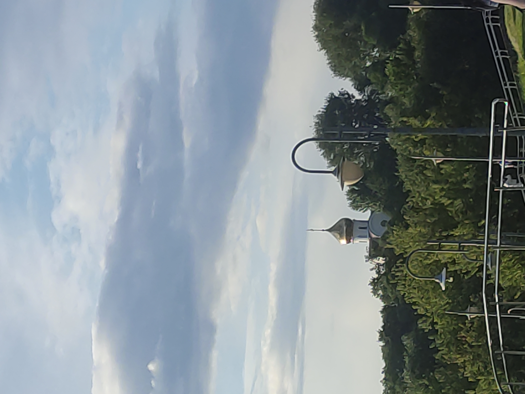 A view of a golden-domed church, nestled amidst lush green trees, is visible under a cloudy sky in Mahilyow, Belarus. A street lamp with a curved metal arm and a light fixture stands in the foreground, with a metal railing to its left. The church's dome shines brightly, reflecting the light from the sky. The trees are dense and form a thick canopy, obscuring the base of the church and creating a natural frame for the religious structure. The sky is filled with soft, white clouds that drift across a pale blue expanse.