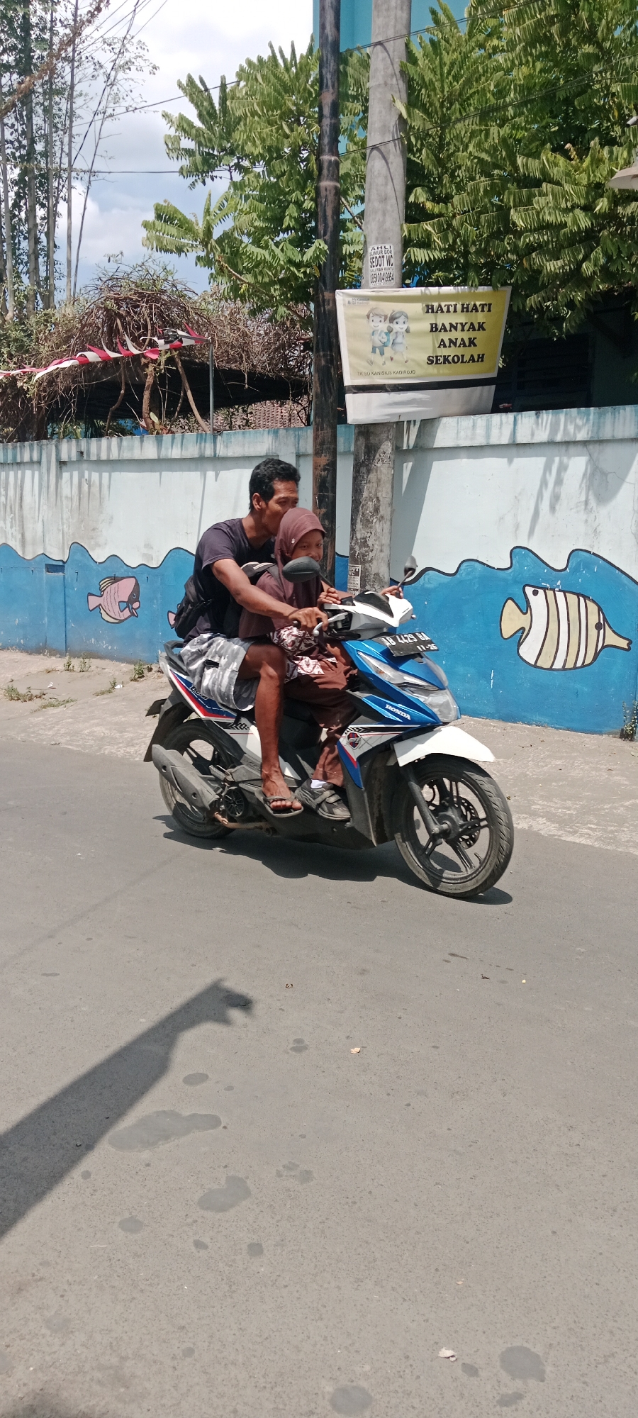 In Purwomartani, Indonesia, on a sunny day, a man and a woman ride a blue and white Honda scooter on a paved road. The man, wearing a dark t-shirt and patterned shorts, is driving. The woman, wearing a brown hijab and patterned dress, is the passenger. Behind them, a blue and white mural of fish adorns a wall. A utility pole stands nearby, with a yellow sign attached that reads "HATI HATI BANYAK ANAK SEKOLAH" (Be careful, many school children). Trees with green leaves are visible behind the wall and pole. The shadow of the scooter and its occupants is cast on the road.