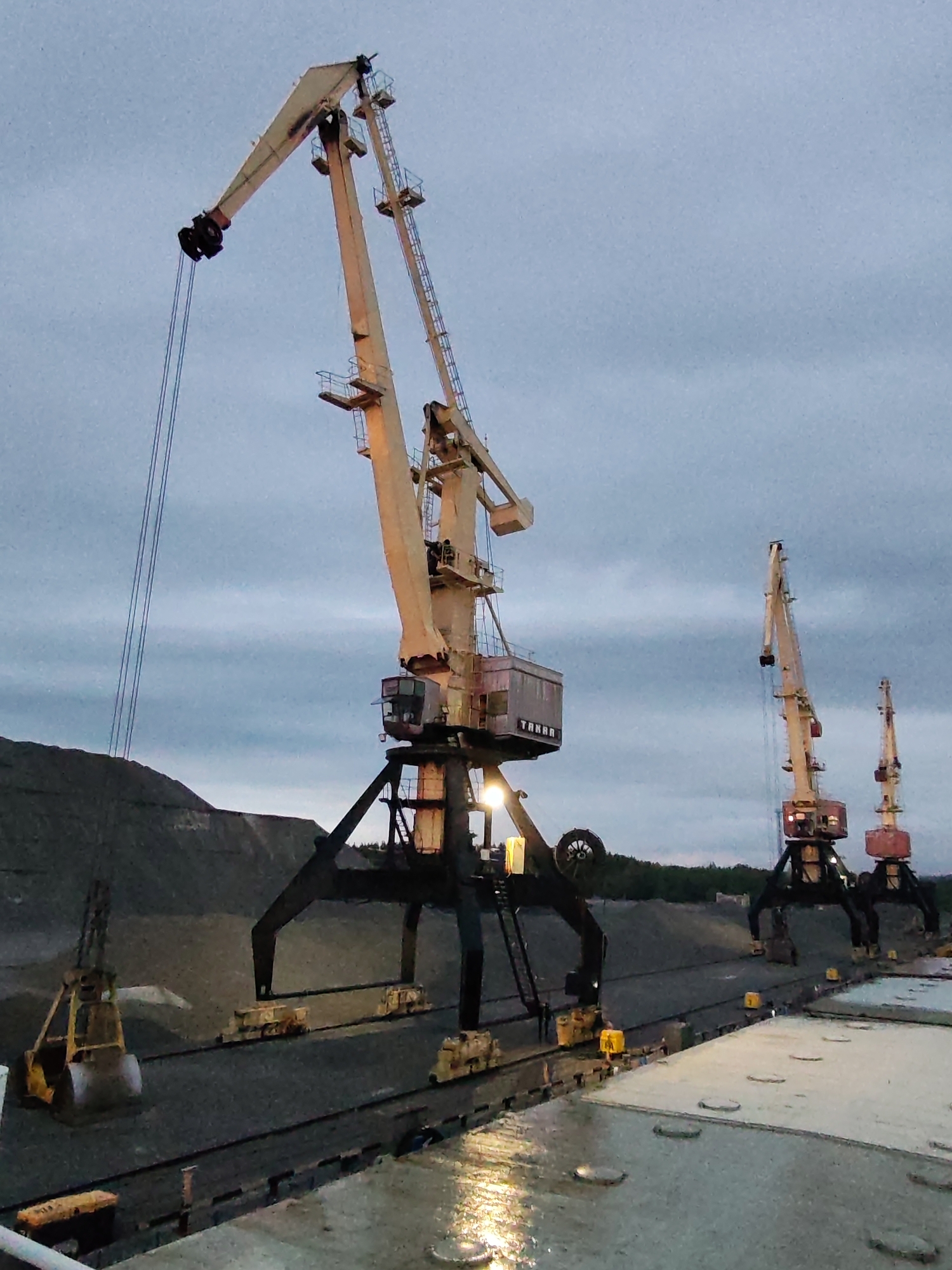 The image shows an industrial scene at what appears to be a port or shipping facility in Shalsky, Russia. Three large, heavy-duty cranes are the dominant feature, with the crane closest to the viewer being the largest and most detailed. The cranes are light in color, possibly beige or pale yellow, and show signs of heavy use.
The nearest crane has its boom extended, and its cables run down to a large scoop. A pile of dark material, possibly coal or ore, is visible behind the scoop and the base of the nearest crane.
The ground is partially paved, wet, and reflective, suggesting recent rain or damp conditions. In the background are trees. The sky is overcast, creating a muted light across the scene.