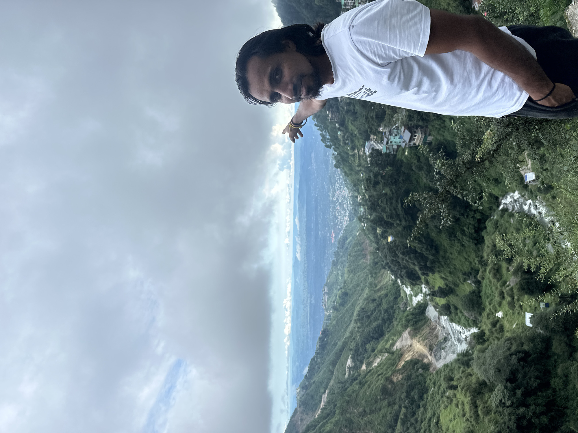 In Bhagsu, India, a man with long dark hair and a beard stands on a hillside, pointing towards a distant valley. He is wearing a white t-shirt and dark pants. The landscape features lush green hills, a river flowing through a valley, and scattered buildings on the slopes. The sky is overcast with heavy clouds.