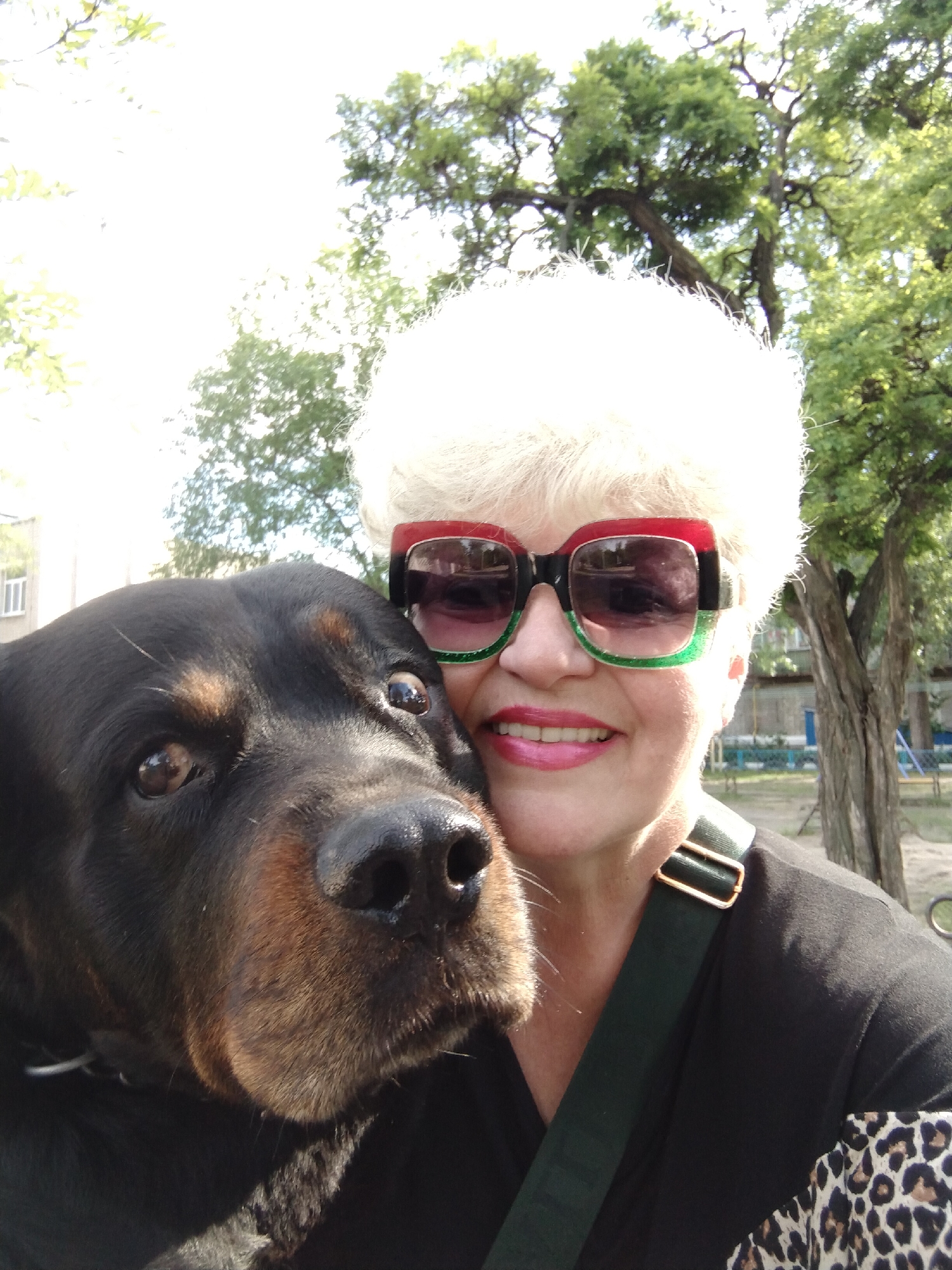 An eye-level, outdoor photograph captures a fair-skinned woman with white, short, curly hair wearing red-and-green-framed sunglasses and a black top with a green strap, posed cheek-to-cheek with a large, black-and-brown Rottweiler. The woman is smiling with bright pink lipstick. The dog's head is pressed against her face, and its eyes are visible.
In the background, there are trees and a glimpse of a light-colored building. The location may be Horishni Plavni, Ukraine, given the prompt.
