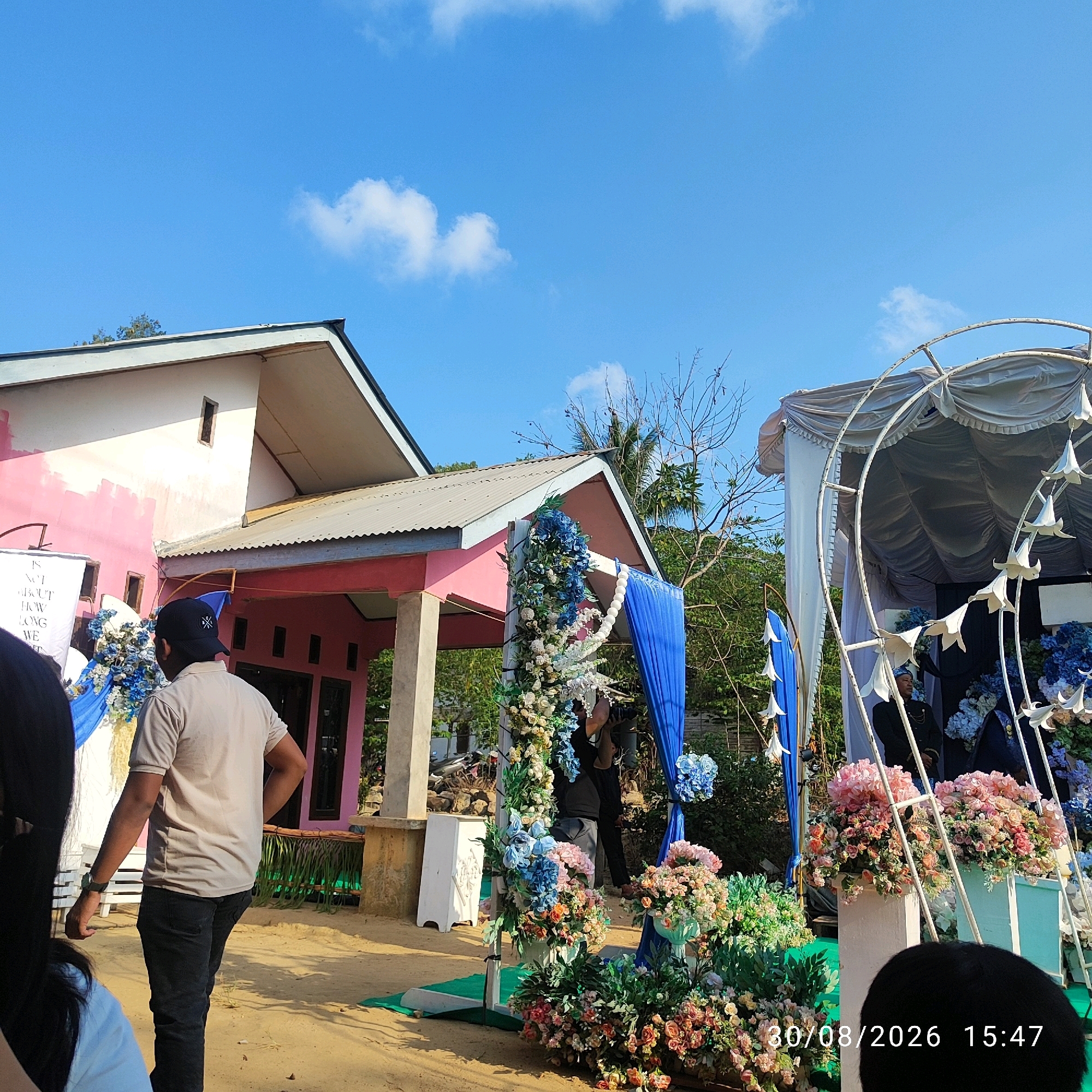 This is a daytime, outdoor, eye-level medium shot photograph taken at a wedding ceremony in Pembaharuan, Indonesia. In the foreground on the left, a person with long dark hair is partially visible, facing away from the camera. To their right, a man wearing a light beige polo shirt and dark pants is walking away from the camera, with his right hand on his hip and a black baseball cap on his head. Behind them, a pink and white building with a silver corrugated metal roof is visible. A white banner with black text is hanging on the left side of the building.
To the right of the building, there is an outdoor wedding decoration setup. This includes an arched white canopy draped with fabric, and a floral archway adorned with numerous blue and white flowers. A blue curtain hangs on the left side of the floral archway. Several white decorative lilies hang from the arched canopy. Two people are visible standing behind the floral archway; one is wearing a dark formal suit and appears to be holding a camera. The ground is sandy and there are various potted plants and floral arrangements around the wedding setup. The sky is blue with scattered white clouds. A timestamp on the bottom right reads "30/08/2026 15:47".