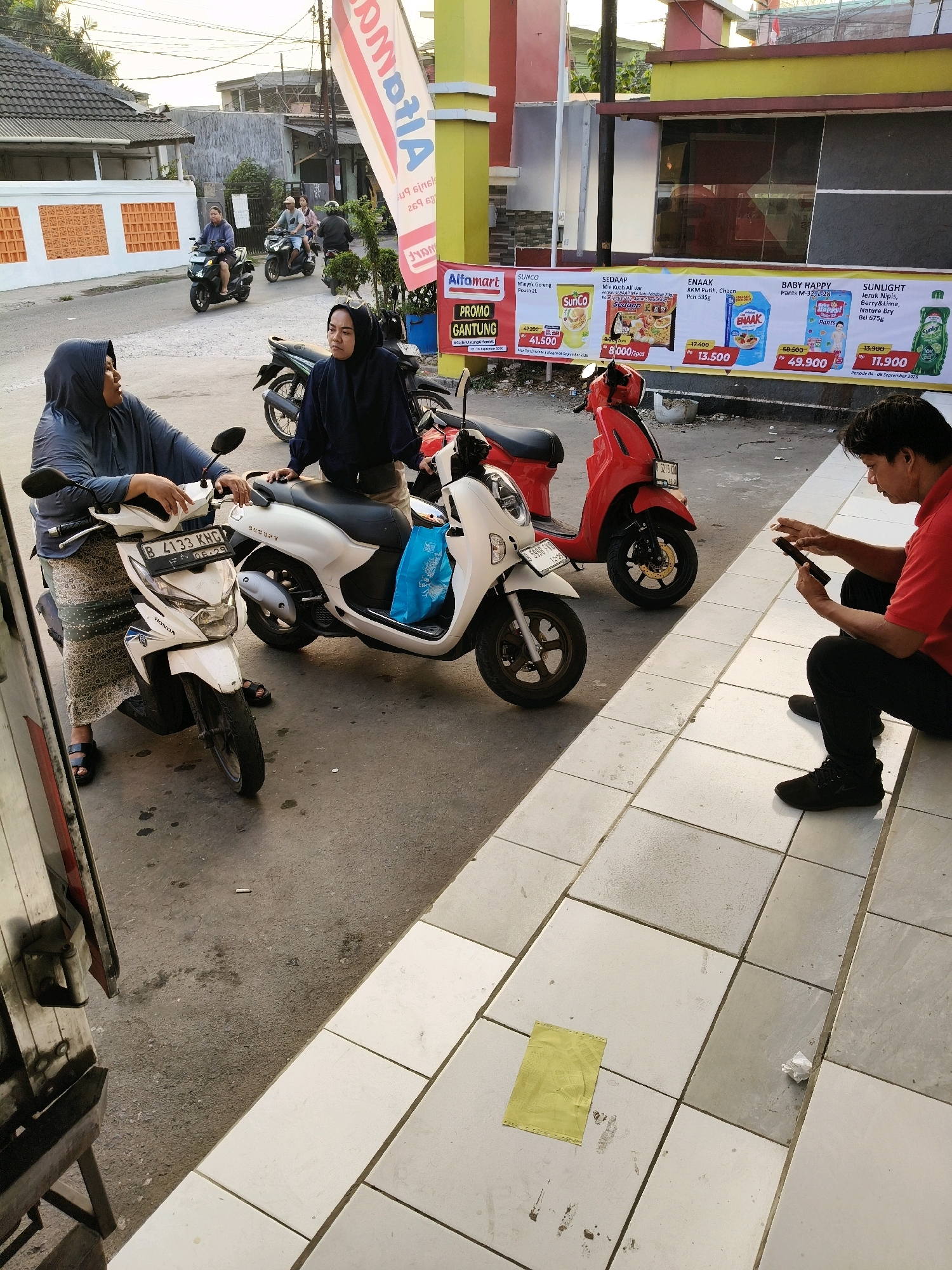 In Setiamekar, Indonesia, two women in hijabs stand near a white scooter, one speaking and gesturing with her hands. A red scooter is parked behind them. Further down the street, several people ride motorcycles, and a man in a red shirt sits on the sidewalk looking at his phone. A banner advertising various products is displayed on the storefront behind the scooters. The ground is paved with concrete tiles, and a yellow piece of paper lies on the ground near the man on the phone.