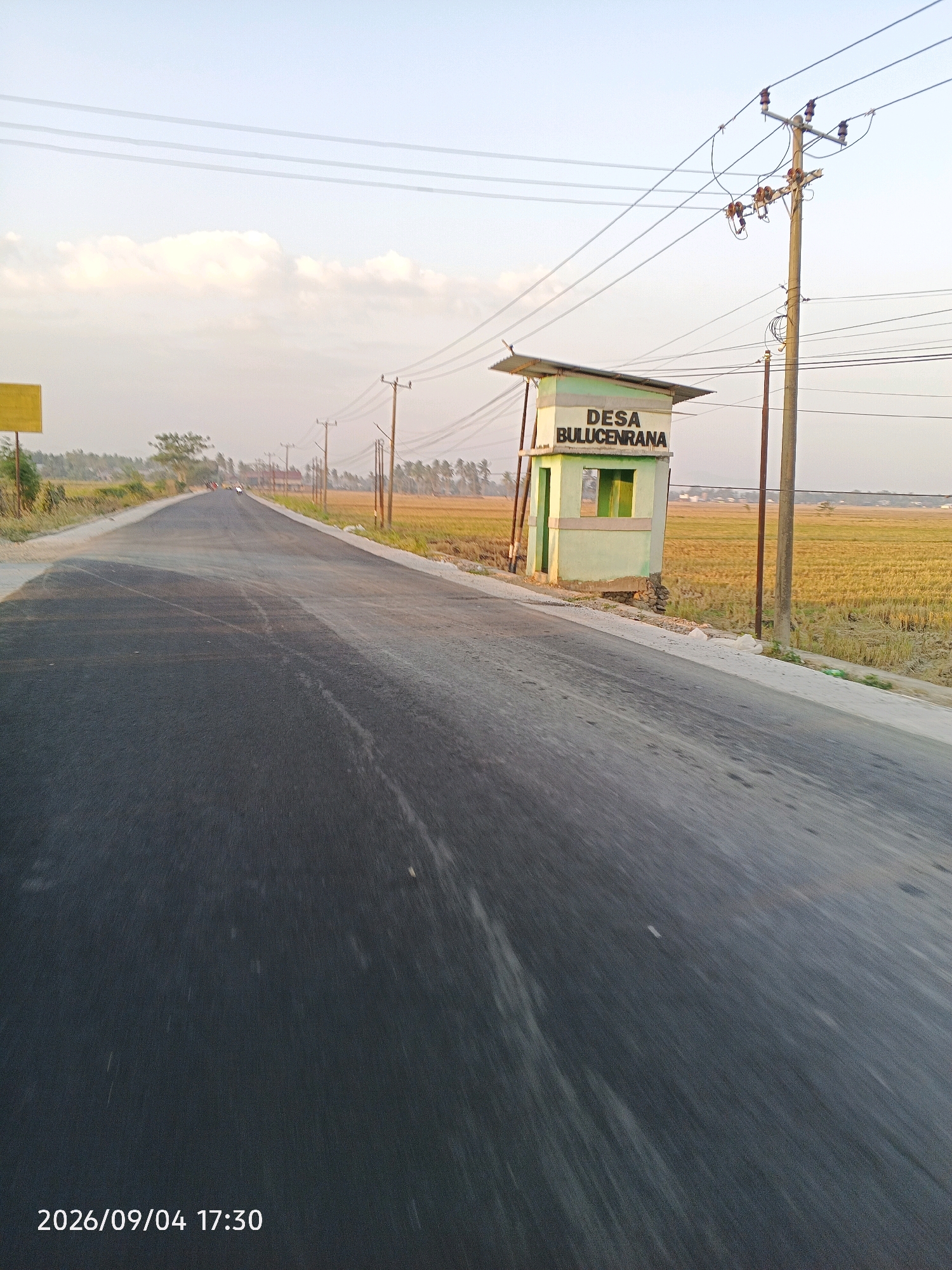 The image shows a straight, paved road in Bulu Cenrana, Indonesia. The road is bordered by dry, harvested rice fields on the right and the left. On the right side of the road, there is a small, light green building with a white roof, possibly a guardhouse or a village information post, with "DESA BULUCENRANA" written in black letters on its side. A utility pole with electrical wires runs parallel to the road on the right. The sky is clear with some scattered clouds, and the lighting suggests it is late afternoon or early evening. A single vehicle is visible in the distance on the road, and there are trees and scattered houses in the background. The timestamp "2026/09/04 17:30" is visible in the lower-left corner of the image.