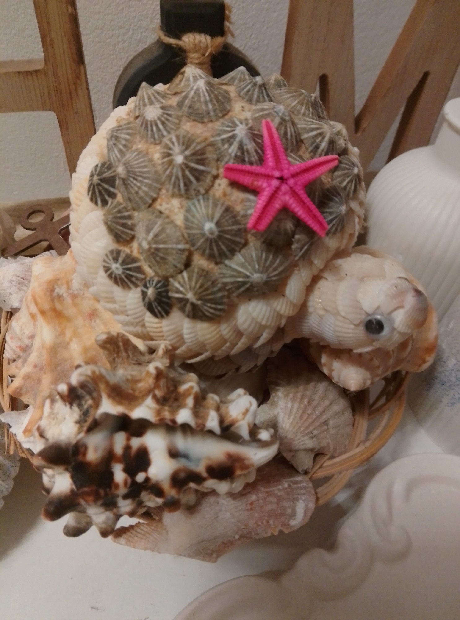 The image displays a decorative object crafted from seashells, resembling a turtle. The turtle's shell is intricately adorned with numerous small, conical shells, and a bright pink, star-shaped shell is prominently placed on top. The turtle's head is also constructed from layered shells, with a dark, circular eye. The entire creation is housed within a woven basket, further embellished with additional seashells. In the background, wooden elements and a white, ribbed object are visible. The scene appears to be an indoor setting, possibly a home, and the image does not provide any specific information about its location in the city of Wedel, Germany.