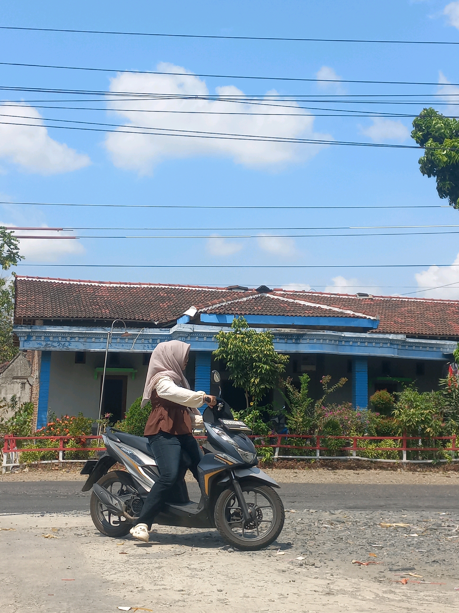 A person wearing a hijab and a brown and white outfit is riding a black and gray scooter on a paved road. The scooter has a license plate that reads "AD 4828 GAE". In the background, a blue and white building with a red tiled roof is visible. There are also trees and bushes along the side of the road. The sky is blue with some white clouds. The scene appears to be in a rural or suburban area of Baturetno, Indonesia.