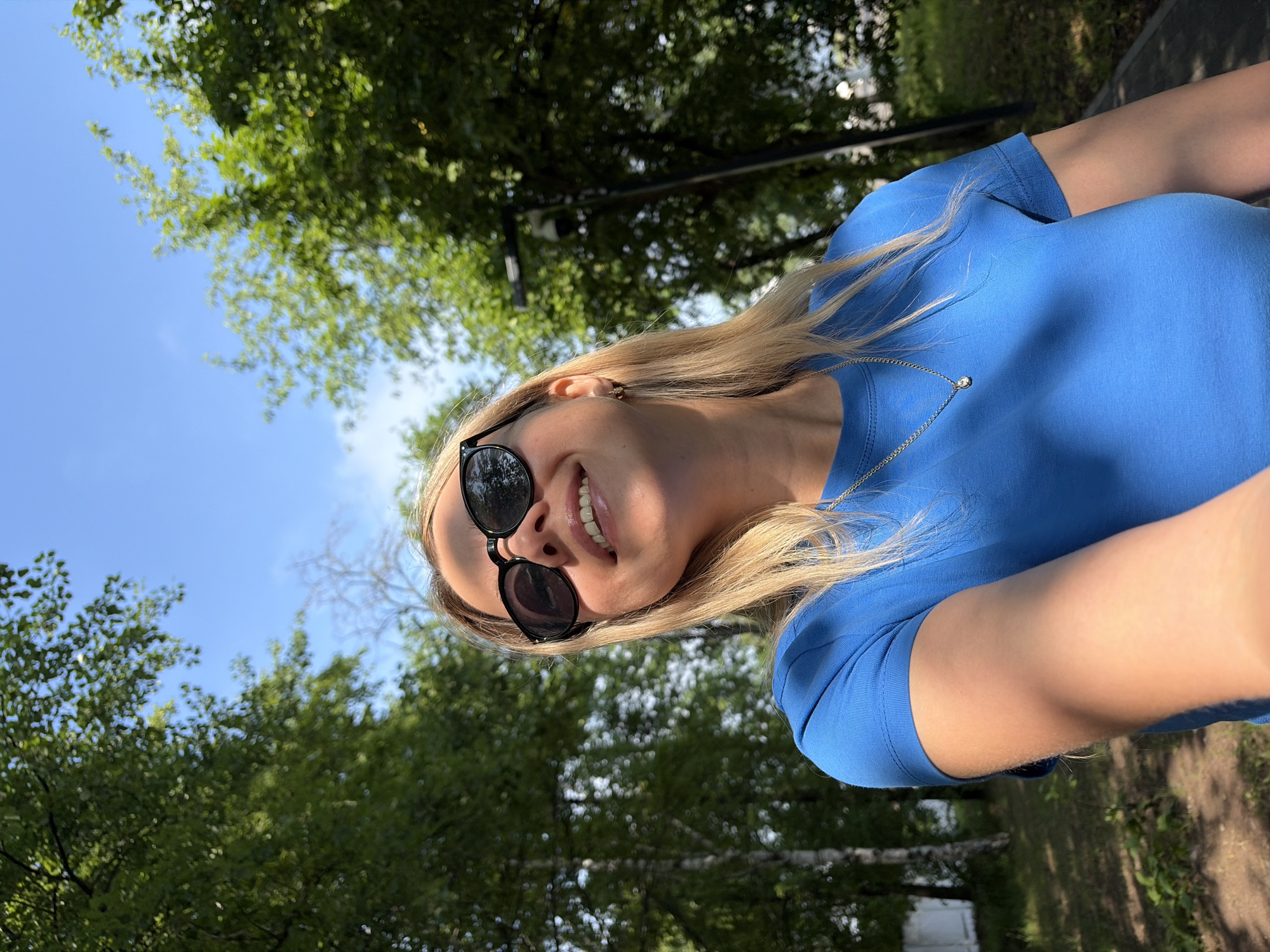 A medium shot captures a blonde woman with sunglasses and a blue t-shirt smiling upwards in a park setting. The background consists of trees with lush green leaves and a clear blue sky, suggesting a sunny day. Sunlight casts shadows on her face and arms. A street lamp is visible in the background. The overall scene appears to be outdoors, likely in a public park within the city of Chaikovsky, Russia.