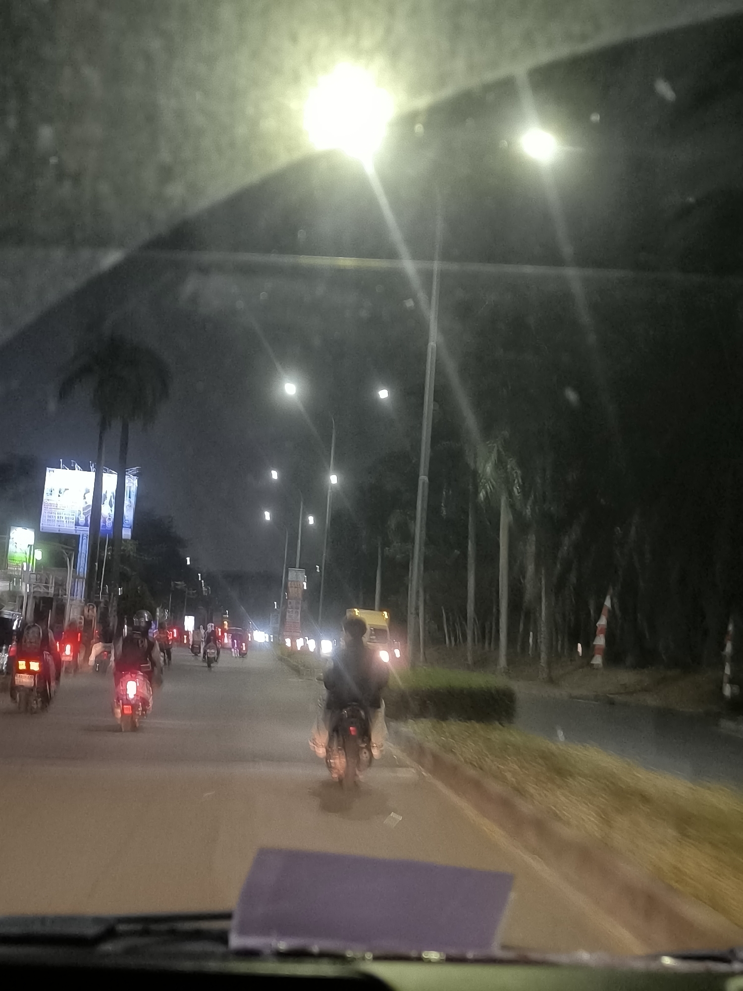 The image captures a nighttime street scene in Tangerang, Indonesia, characterized by heavy traffic, primarily consisting of motorcycles. The perspective is from inside a vehicle, looking out through a windshield, which is partially obscured by a light purple glare in the foreground.
The road is illuminated by numerous streetlights, some of which are bright and cause glare. Several palm trees are visible along the roadside, some lining the street and others forming a denser cluster in the background, indicating a tropical or subtropical environment.
The traffic flow includes a significant number of motorcycles with their taillights illuminated, suggesting they are moving or stopped in traffic. Some of these motorcycles have riders visible. There are also some cars and possibly a bus or truck present in the scene, identifiable by their headlights and taillights.
In the background, there are buildings and billboards, one of which is brightly lit and displays advertisements, suggesting an urban or commercial area. The overall atmosphere is that of a busy city street at night, with vehicles and pedestrians navigating through the darkness. The image is somewhat blurry due to motion or low light conditions, but the essential elements of the scene are discernible.
