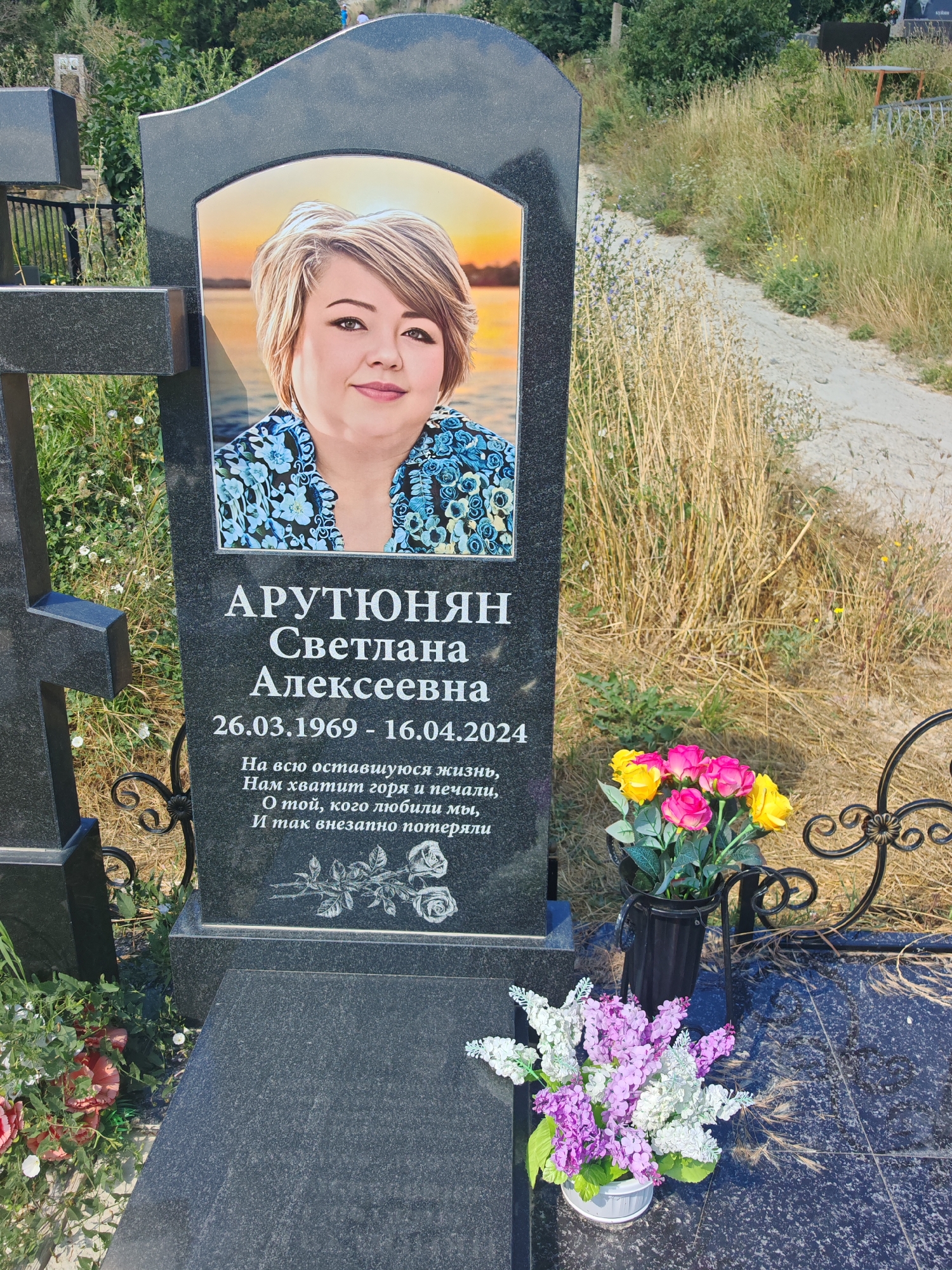 The image shows a black granite headstone with a portrait of a woman and an inscription. The woman in the portrait has blonde hair and is wearing a blue floral shirt. The inscription includes the name "АРЮТЮНЯН Светлана Алексеевна" and dates "26.03.1969 - 16.04.2024". Below the dates, there is text in Russian, which translates to "For all the remaining life, we will not have enough grief and sorrow. For you, whom we loved, and lost so suddenly." There is also an etched image of roses on the headstone. Two vases of artificial flowers, one with pink and yellow roses and another with purple and white flowers, are placed in front of the headstone. The headstone is located in a cemetery with dry grass and some greenery in the background, suggesting an outdoor setting. The overall scene is of a grave memorial.