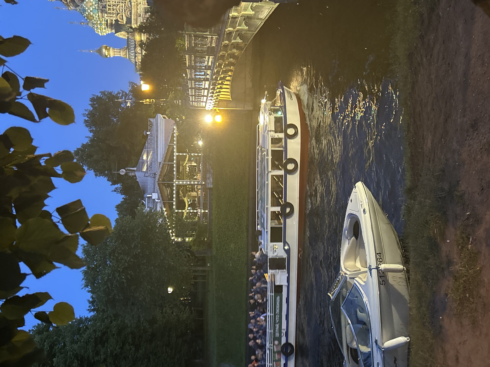 This is a nighttime medium shot of a canal or river in an urban setting, possibly St. Petersburg, Russia, given the architectural style of the illuminated domed building in the background, likely the Church of the Savior on Spilled Blood. In the foreground, a small white motorboat with the registration number "51264" is moored at the grassy bank. Further back, a larger white and blue tour boat carrying numerous passengers travels down the waterway. The tour boat has text in Russian on its side, "ДЕЛАЙ, ЧТО ЛЮБИШЬ." The water reflects the lights from a bridge, a lamppost, and the buildings. Trees line the embankment. The sky is a deep blue, indicating it is dusk or night.