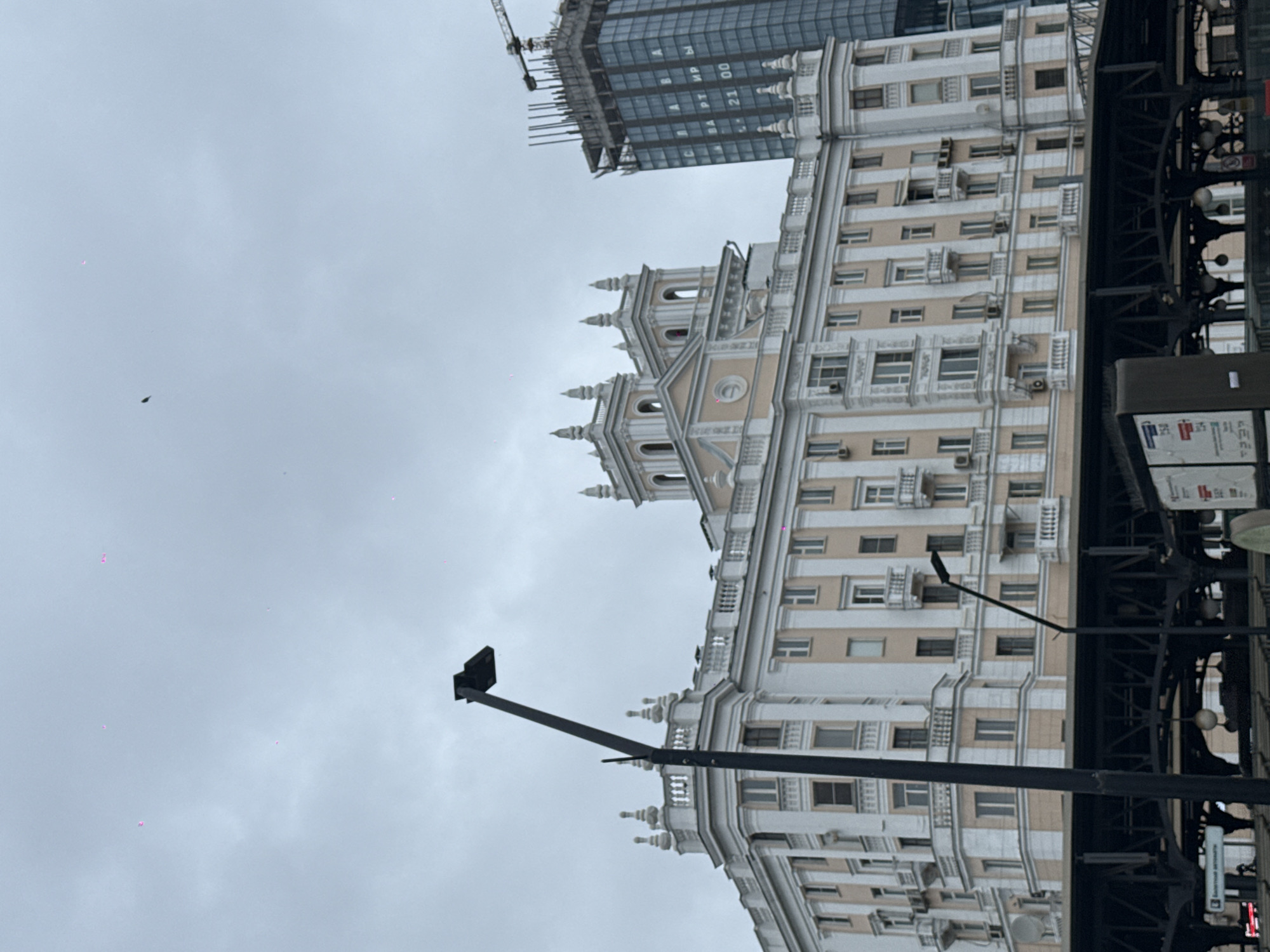 This is a vertical, wide-angle photograph of an urban landscape in Moscow, Russia, taken on an overcast day. The scene captures a juxtaposition of architectural styles under a grey, cloudy sky.
In the midground, a stately, multi-story building with a light-colored facade is prominently featured. It exhibits ornate architectural details, including decorative balustrades, cornices, and numerous windows, with many air conditioning units mounted on its exterior. This building appears to be in a classical or neoclassical style.
Behind and to the left of this ornate structure, a modern, dark glass-clad skyscraper is under construction. A large construction crane is visible at its apex, indicating ongoing development. Text in Cyrillic letters is visible on the modern building's facade, spelling out "КВАРТИРЫ" (Apartments) and "21 00."
In the foreground, a dark, heavy metal structure, possibly part of an elevated walkway or bridge, partially frames the right side of the ornate building. A black street light pole with a rectangular fixture extends from the lower right into the middle of the frame. Below the metal structure, a sign with the Cyrillic word "БАНКОМАТЫ" (ATMs) is partially visible.
The sky is a uniform light grey, suggesting overcast weather. A few faint, small pink artifacts, likely sensor dust, are scattered across the sky. No people are clearly visible, and the overall impression is that of a bustling urban environment characterized by both historical presence and ongoing modern development in a large city.